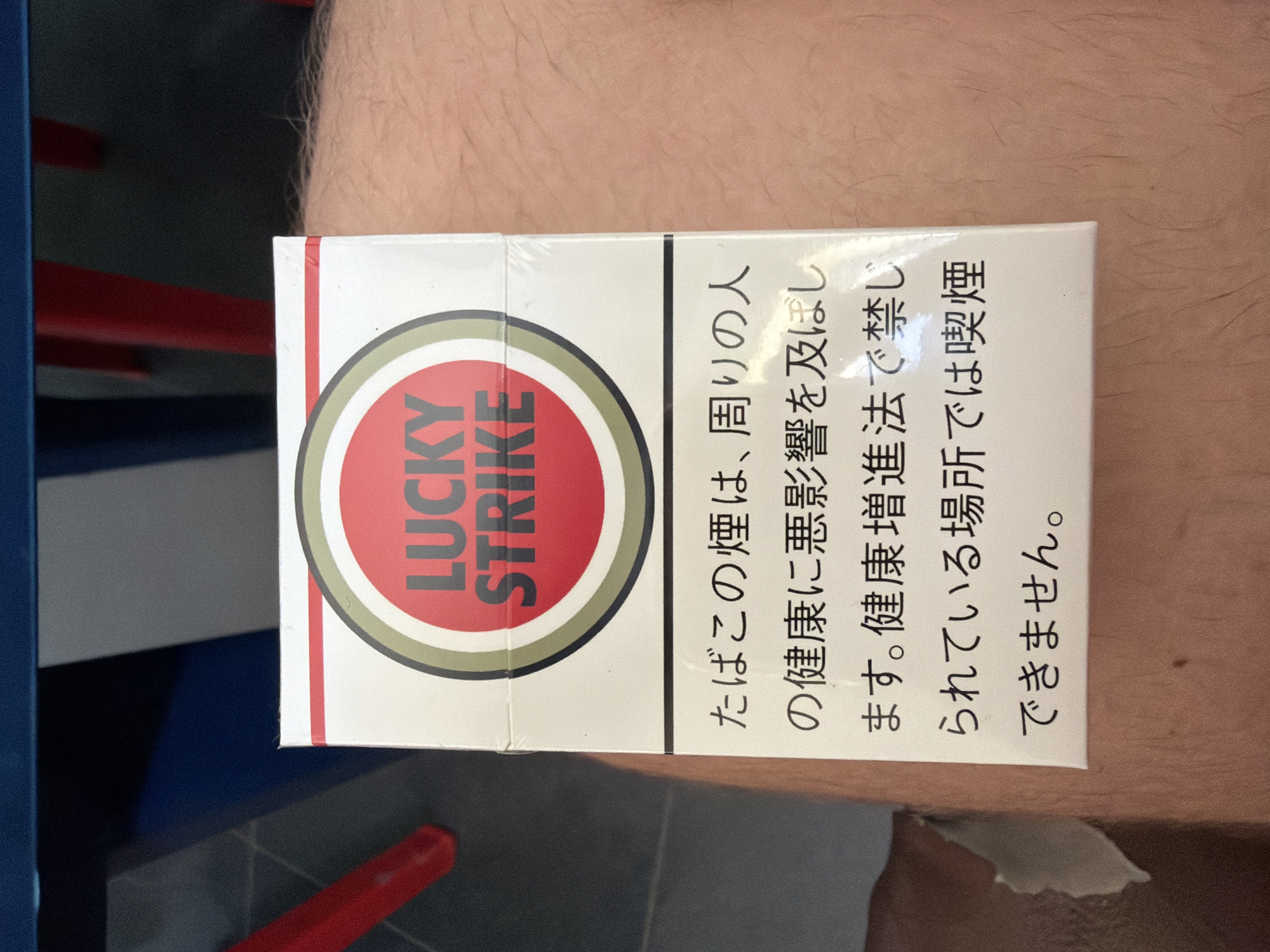 An overhead shot shows a new, cellophane-wrapped pack of Lucky Strike cigarettes resting on a person's light-skinned, hairy leg or arm. The pack is oriented vertically and rotated 90 degrees clockwise, with the classic Lucky Strike logo—a red circle with "LUCKY STRIKE" in white, bordered by black and olive green rings—prominently displayed on a white background. To the right of the logo, Japanese text is visible, which reads: "たばこの煙は、周りの人の健康に悪影響を及ぼします。健康増進法で禁じられている場所では喫煙できません。" This translates to: "Tobacco smoke adversely affects the health of those around you. Smoking is prohibited in places where it is forbidden by the Health Promotion Act." In the out-of-focus background, dark shapes suggest an indoor setting, with hints of a blue surface on the left and a red object below. The time of day is not discernible. The image provides a close-up view of the cigarette pack against a personal backdrop, likely taken in Chervena voda, Bulgaria.