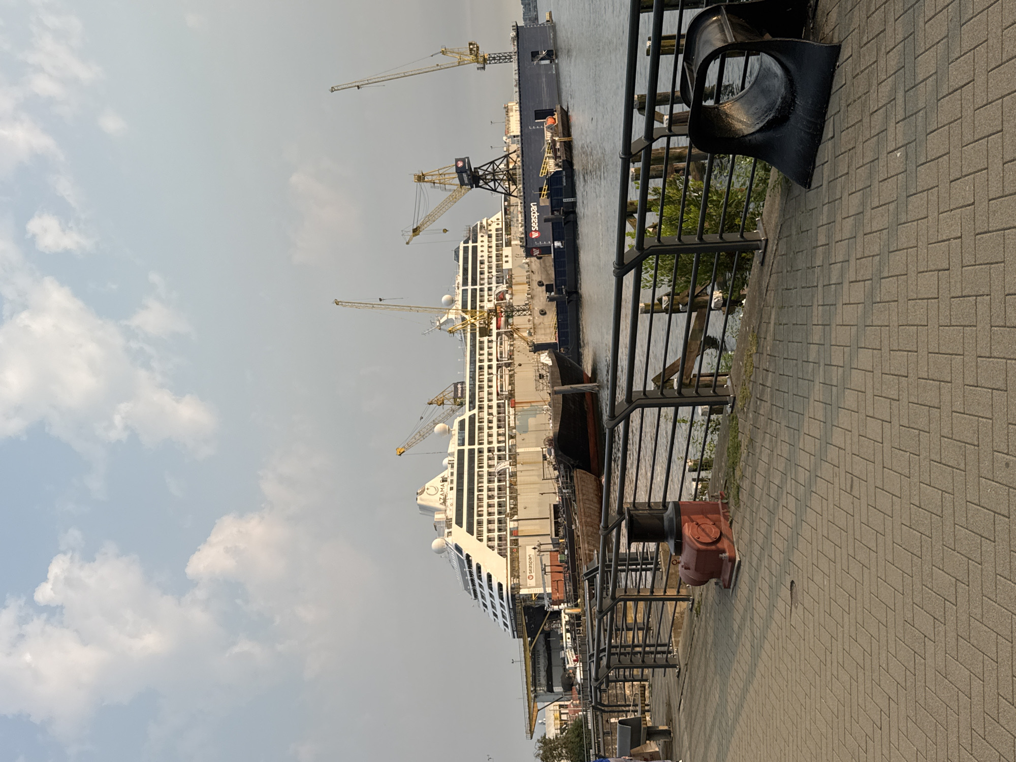 In the city of North Vancouver, Canada, a large white cruise ship, identified as the "AZAMARA PURSUIT," is docked in a dry dock. The ship is surrounded by industrial elements, including a dark blue floating dry dock structure and several yellow cranes. The scene depicts a shipyard environment. In the foreground, a paved walkway is visible, bordered by a black metal railing. A red industrial bollard is present on the walkway, and to its right, a large black curved object is situated. The water of the harbor is visible beyond the railing. The sky above is partly cloudy, with the sun casting a warm light on the scene. There are no visible people in the immediate foreground, but there are some distant figures on the ship. The overall atmosphere is one of maritime industry and activity.