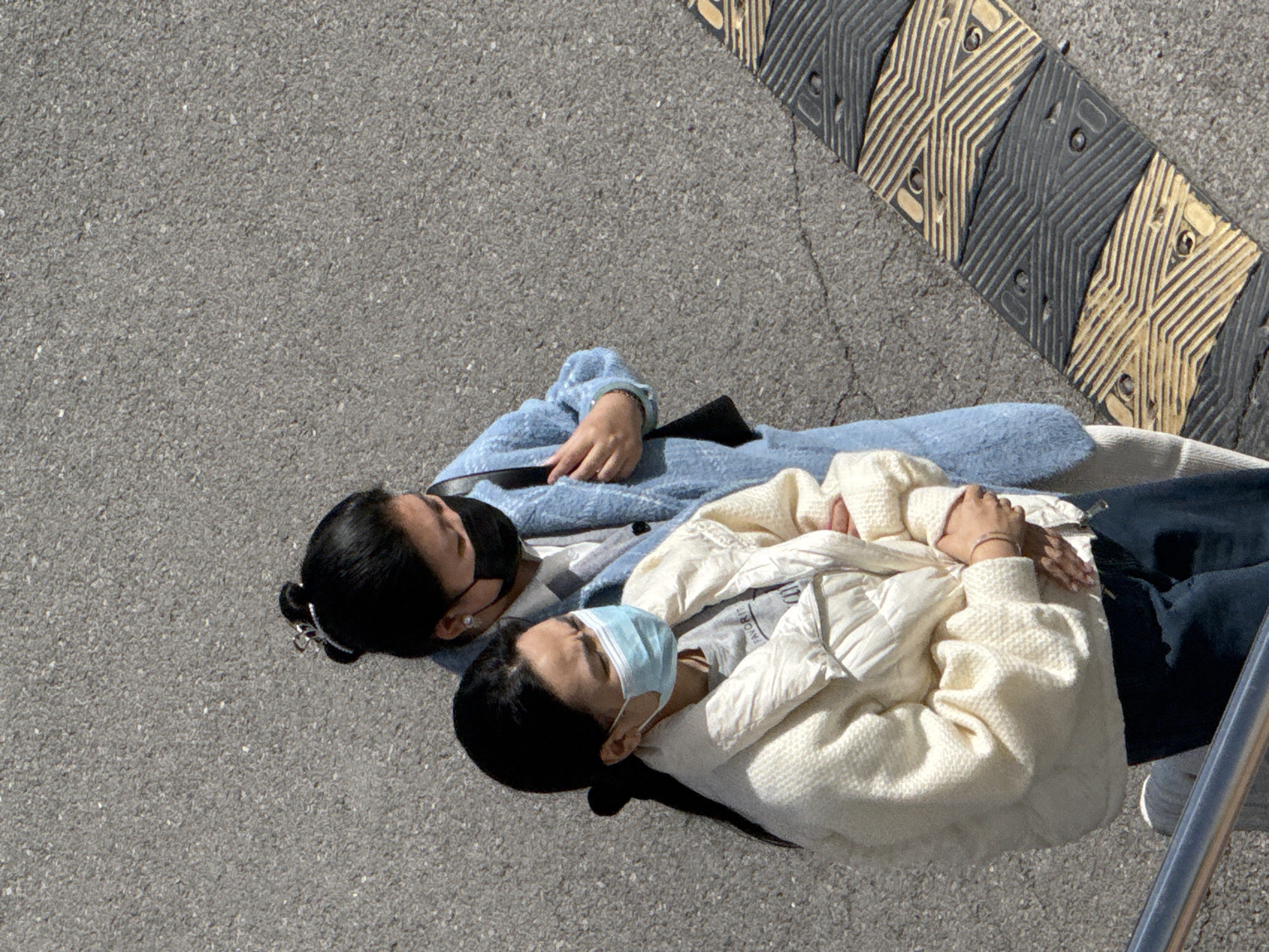 Two people are lying on an asphalt surface next to a yellow and black speed bump, presumably in the city of Zhongtun, China. The person closer to the speed bump is wearing a white puffer jacket, a gray shirt, dark pants, and white shoes and has a light blue disposable face mask covering their nose and mouth. The other person is wearing a light blue cardigan over a white top, dark pants, a black shoulder bag, and a black face mask. The faces of both individuals are visible. The surrounding environment is a gray asphalt surface.
