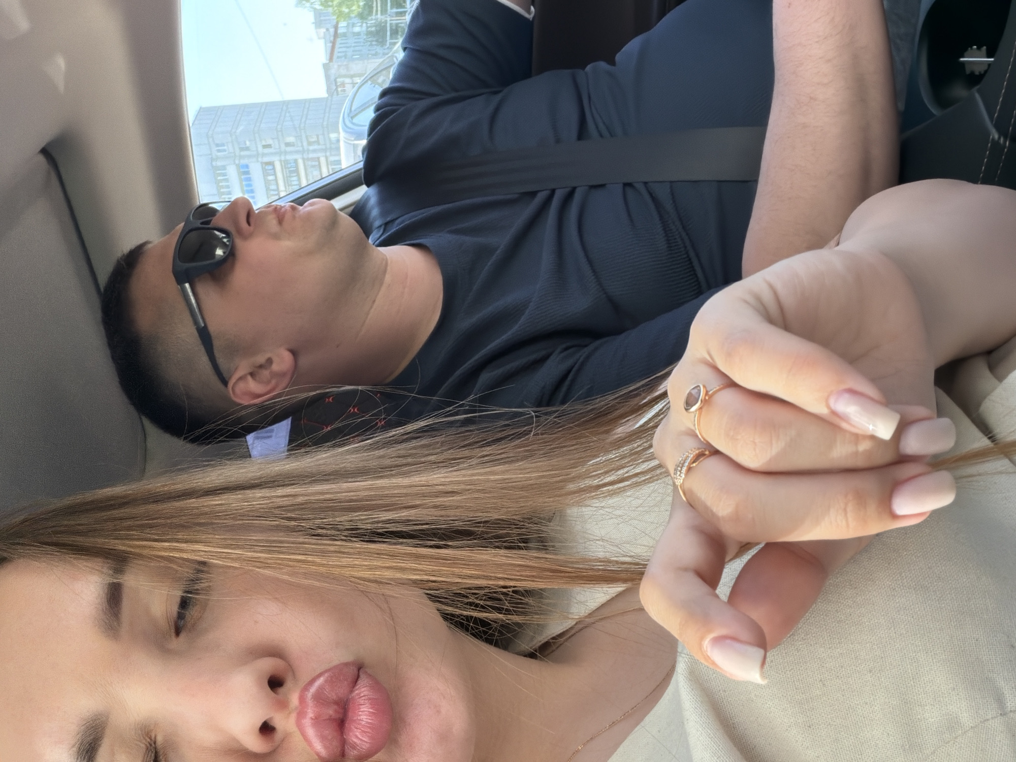 The image depicts a couple taking a selfie inside a car, possibly during a ride in Chaikovsky, Russia. The man, wearing sunglasses and a dark-colored shirt with a seatbelt, appears to be leaning back in the passenger seat with his eyes closed. The woman, presumably in the driver's seat, is taking the selfie, her face partially visible with pursed lips and long blonde hair. Her hand, adorned with two rings, is in the foreground. Outside the car window, buildings can be seen, suggesting an urban environment.