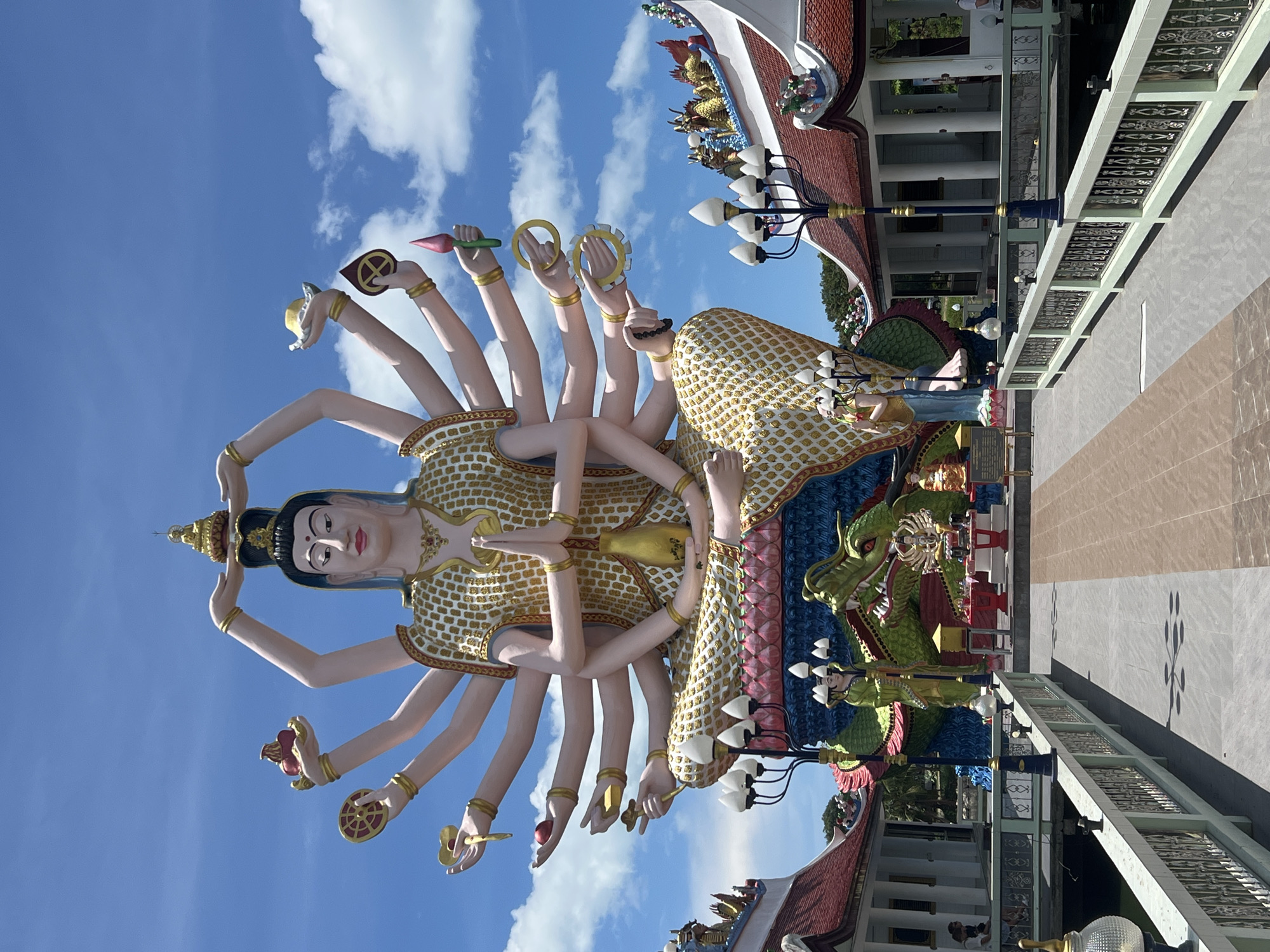 The image depicts a statue of Guan Yin with eighteen arms, a significant landmark in Baan Plai Laem, Thailand. The statue stands tall under a bright blue sky dotted with white clouds. The statue's complexion is fair, and the intricate patterns on its clothing are painted in gold and white. Each of its eighteen arms holds a different object, including a lotus flower and a wheel. Below the statue, there is a green dragon statue, and behind it, a building with a red roof and white columns. The area around the statue is paved with light gray tiles, and a decorative metal railing can be seen. A row of streetlights with white globes is situated near the statue.