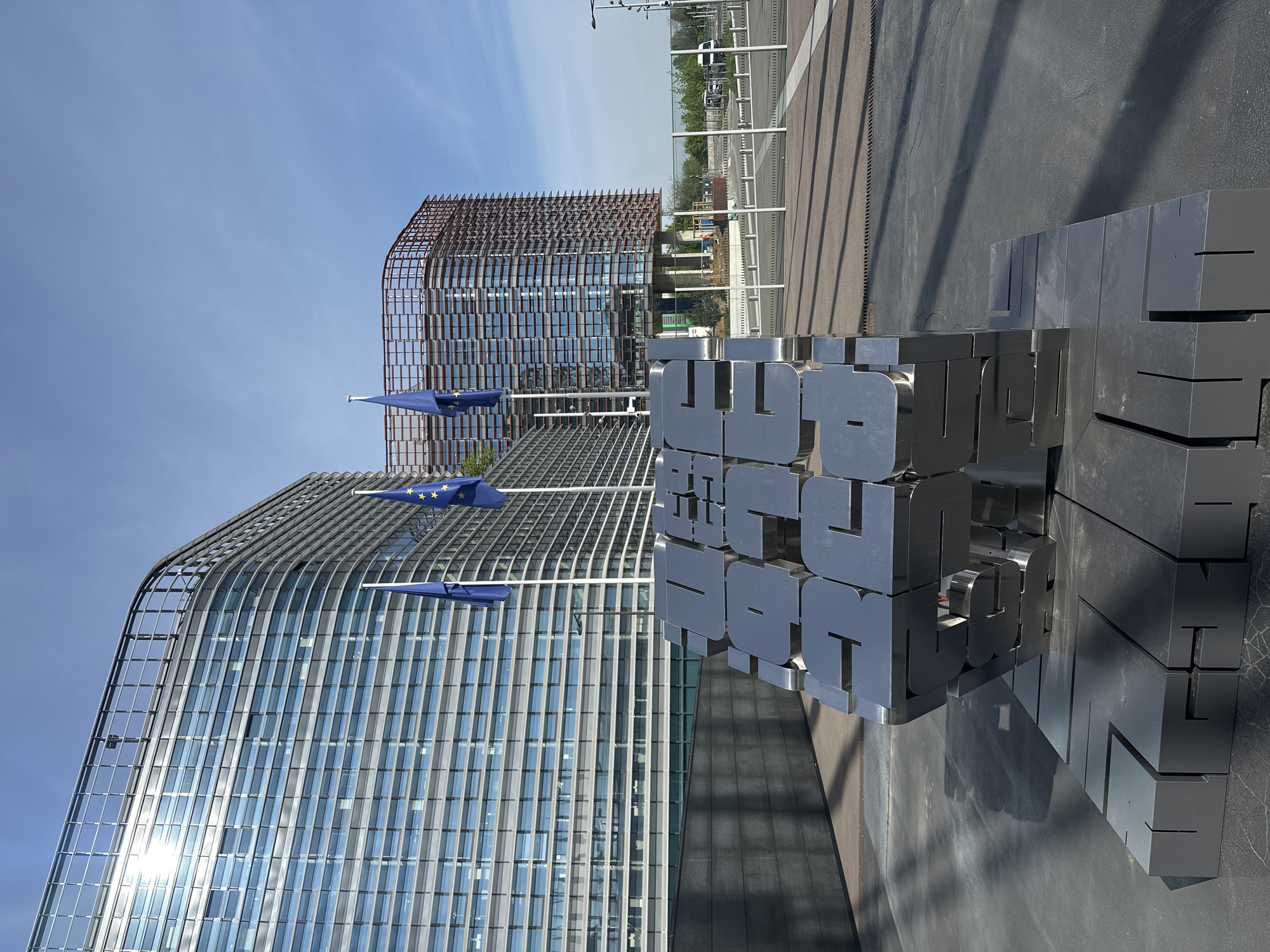 This outdoor image, taken in Strasbourg, France, depicts a modern architectural landscape under a clear blue sky. In the foreground, a large, metallic, cube-shaped sculpture dominates, composed of block-like letters. The sculpture clearly spells out "EUROPEAN PARLIAMENT" on its front face, and partially "EUROPEEN PARLEMENT" on its right side, identifying the location's institutional context.
Behind the sculpture and to the left, a sleek, modern building with a curved glass facade reflects the bright sunlight. In front of this building, three European Union flags, blue with yellow stars, fly from poles. Further back, another contemporary building is visible, featuring a distinctive facade of reddish-brown lattice work or panels.
The scene is bathed in bright daylight, casting defined shadows, suggesting a sunny mid-morning or afternoon. The ground is paved, resembling a plaza or pedestrian area, with a road or parking lot visible in the background, separated by white barriers. The overall atmosphere is one of governmental modernity and European identity, with no specific human activity taking place.