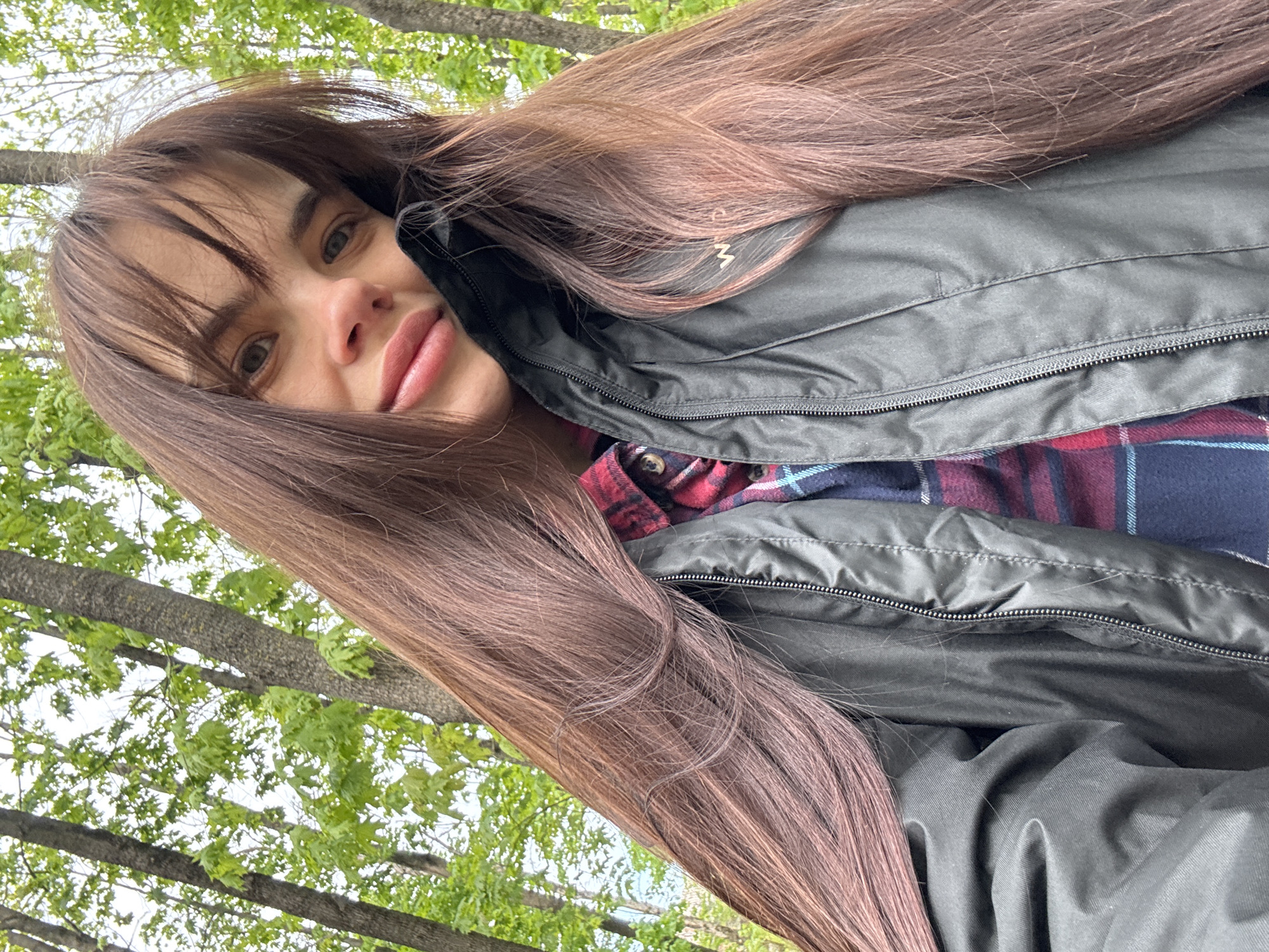 A woman with long, straight, light brown hair and bangs is posing for a selfie outdoors. She has fair skin, light eyes, and full lips. She is wearing a dark gray, quilted jacket over a red and blue plaid shirt. The background includes trees with green leaves. The location is not discernible as Rostov-on-Don or any other specific place.