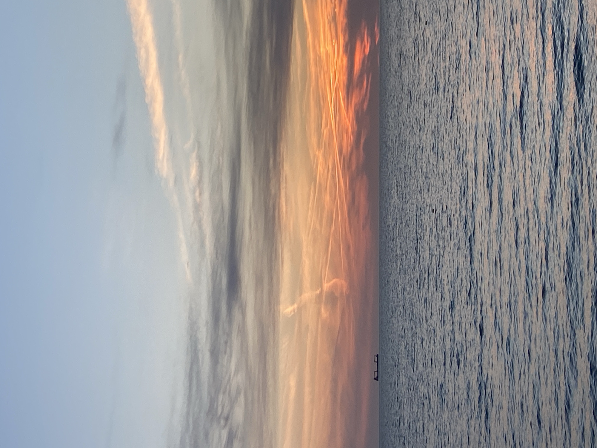 The image captures a sunset over the Adriatic Sea near Bar, Montenegro. The sky transitions from a pale blue to orange and red near the horizon, marked with contrails. A body of water is visible on the right, with a small dark silhouette of a ship on the horizon. The water surface reflects the sunset's colors.