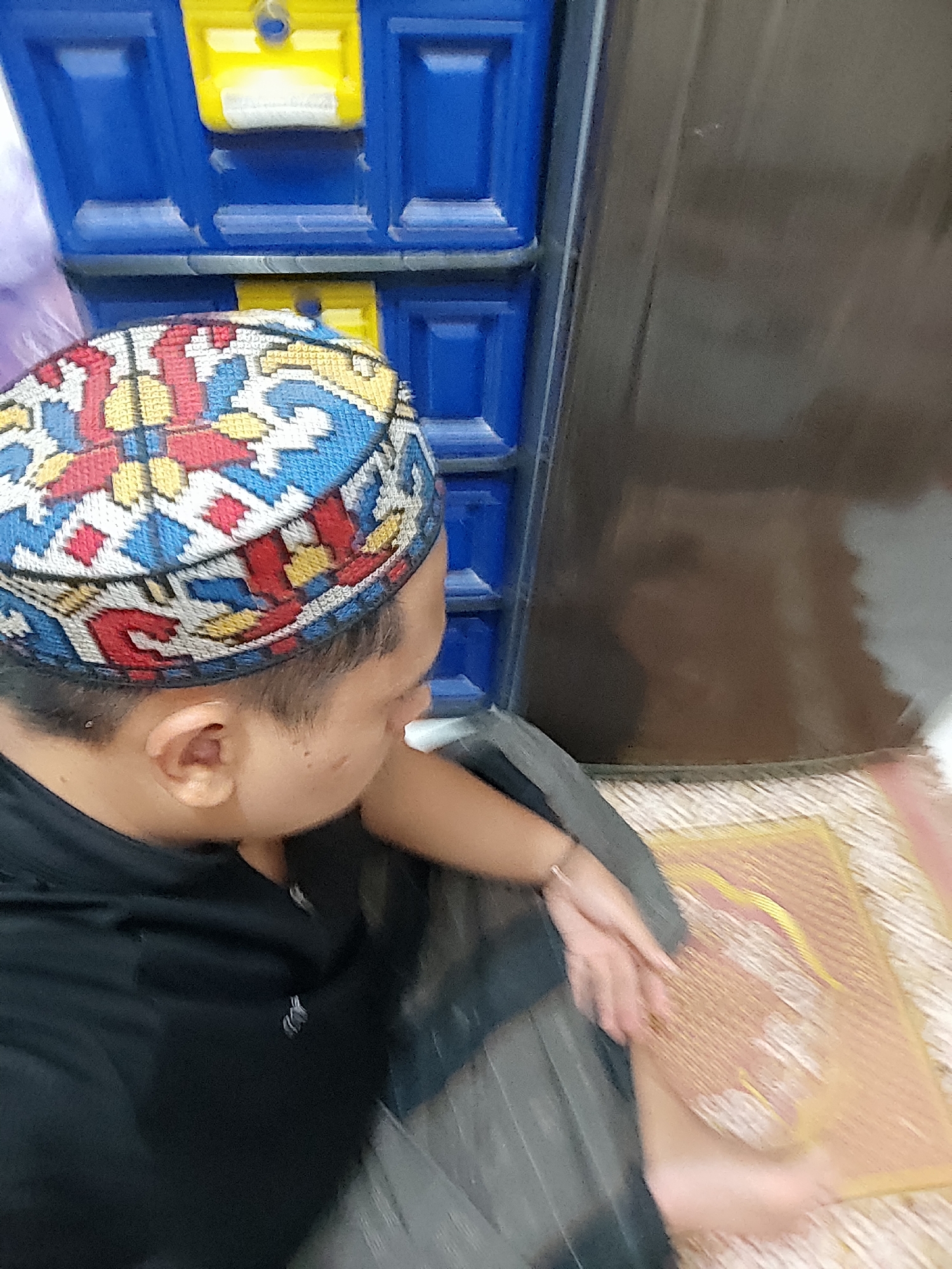 A person is shown from the shoulders up, wearing a black shirt and a kufi with a colorful, patterned design. The kufi is red, blue, and yellow, with geometric shapes. The person's face is not fully visible, as they are looking down and to the right. In the background, there is a blue storage unit with yellow drawers. To the right of the person, a section of a dark brown, possibly wooden, surface is visible. The person's right hand is resting on a textured surface that appears to be a mat with embedded pins. The flooring is a woven mat. The scene suggests an indoor setting, possibly a home. The location in Satriamekar, Indonesia is not explicitly visible in the image.
