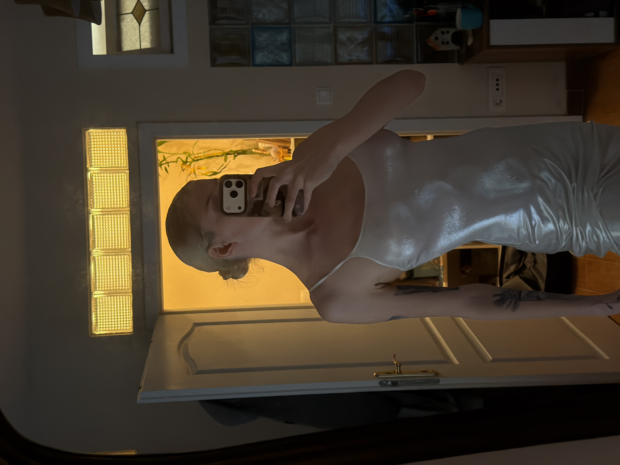 A person is depicted taking a mirror selfie indoors. The subject, likely female, has light-colored hair styled in a bun and holds a smartphone with a black and white case, which partially covers their face, obscuring the eyes and nose. They are wearing a light-colored, shiny, form-fitting slip dress or nightgown. A tattoo featuring several palm trees is visible on their right upper arm.
The reflected background shows an interior space illuminated by warm, artificial light. To the left, a vertical stack of three textured glass blocks glows brightly with a yellow light. Next to these, a white door frame surrounds a slightly ajar door, revealing a brightly lit area inside with green foliage, possibly bamboo, visible. Above the door frame, a row of horizontal glass blocks, some appearing with a blueish tint, are set into the wall. Further right, a white wall-mounted power outlet with three sockets is visible. Above this, shelves or furniture hold various items, including a light blue object. The floor in the lower right of the reflection appears to be tiled.
The warm artificial lighting suggests the photo was taken during the evening or night, or in a room relying on internal illumination. There is no visible text in the image. The location context provided is Petrovac na Moru, Montenegro.
