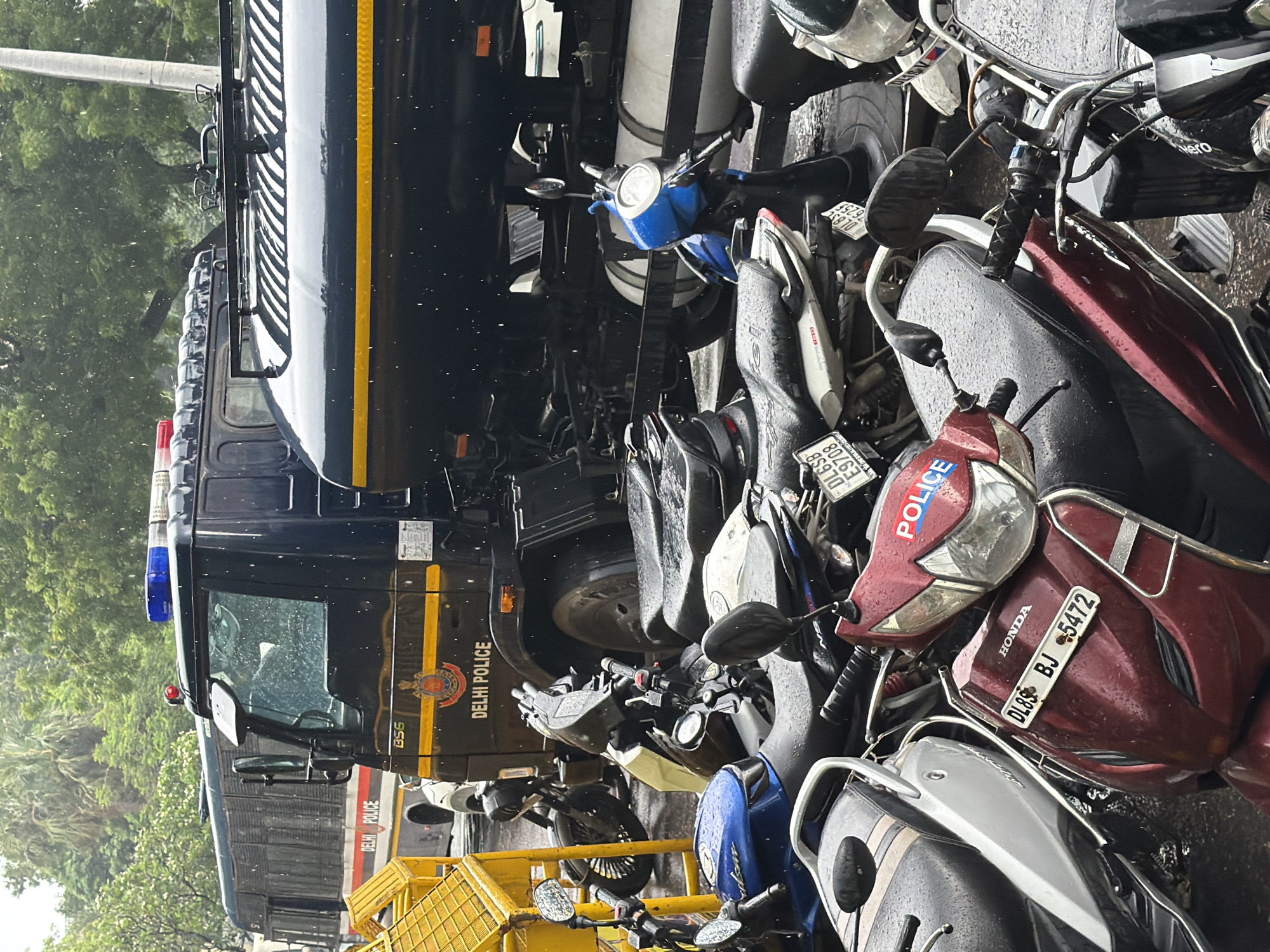 The image depicts a scene in New Delhi, India, featuring multiple parked motorcycles and a large Delhi Police truck. It appears to be raining, with visible raindrops and wet surfaces. The Delhi Police truck, dark blue with yellow and red accents, has "DELHI POLICE" written on its side, along with a police emblem. Its roof is equipped with blue and red flashing lights. Next to the truck, a yellow barrier is visible. Several motorcycles are parked in front of the truck, some in close proximity. One motorcycle in the foreground is maroon with "HONDA" and a license plate "DL8S BJ 5472" clearly visible. Another motorcycle, red with white lettering "POLICE", is also prominent. The overall environment suggests a street or parking area in an urban setting. There are no people visible in the image.