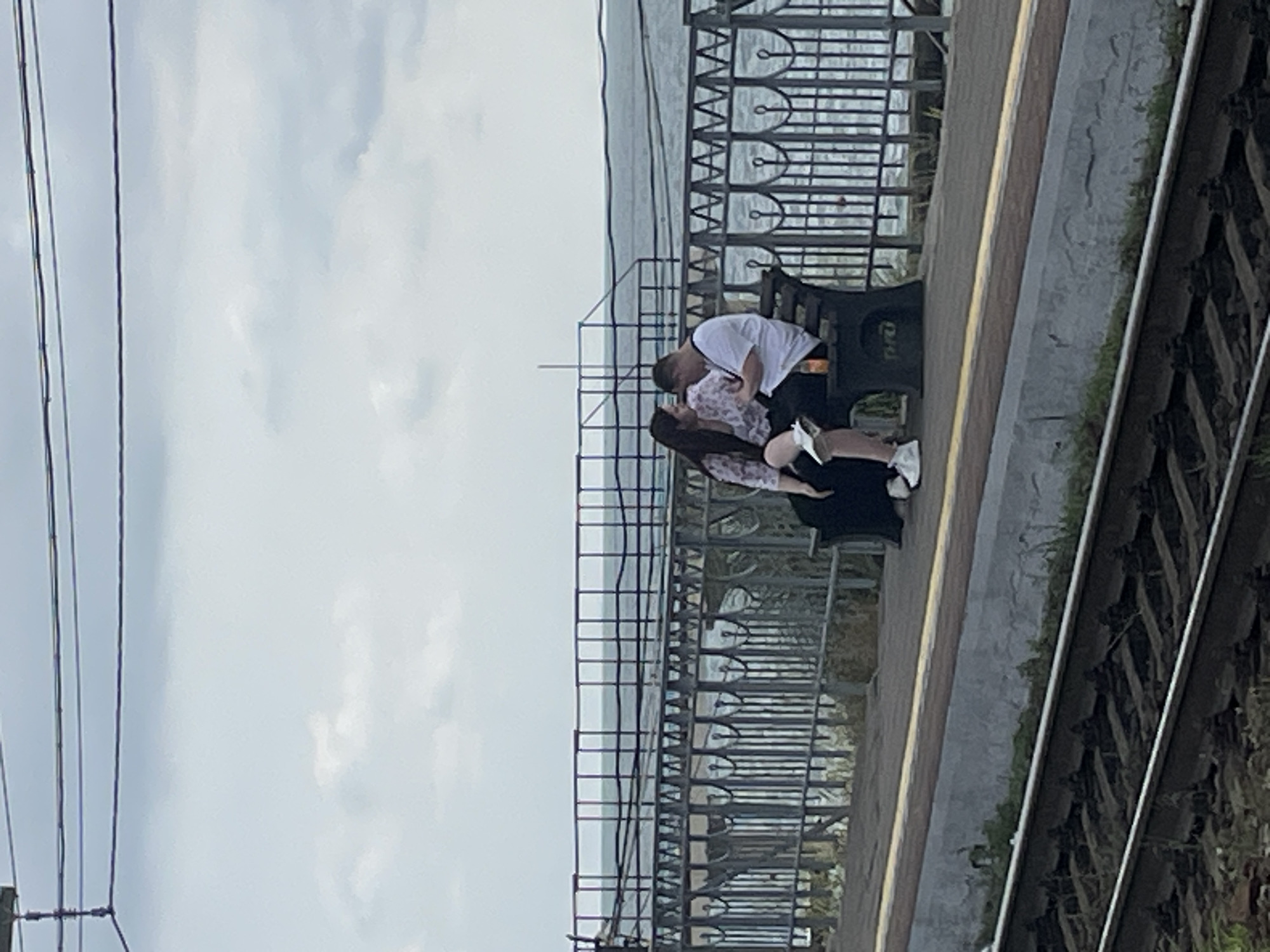 Two individuals, a man and a woman, are seated on a dark-colored bench on a promenade. The woman, on the left, has long dark hair and is wearing a light-colored floral-patterned top and dark pants. The man, on the right, is wearing a white t-shirt and dark shorts. He appears to be looking down at something in his hands. There is a bottle on the bench to his right. They are situated on a promenade overlooking the sea, with a metal fence separating them from the water. In the foreground, train tracks are visible. The sky is overcast. The scene suggests a location in Sochi, Russia, possibly a train station or a coastal walkway.