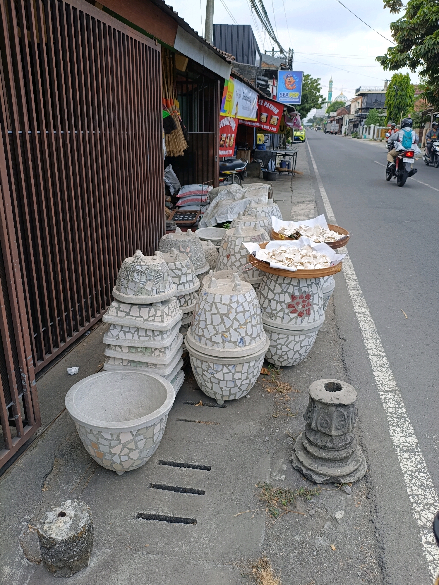 The image shows a street scene in Purwomartani, Indonesia. In the foreground, on the sidewalk, are several concrete pots, some stacked and decorated with mosaic tiles. One large, empty mosaic pot sits on the ground. Next to it is a short, weathered concrete post with a hole in the top. A second, more ornate, broken concrete post stands near the edge of the sidewalk. In the middle ground, a row of shops line the street, with brooms hanging outside one of them. Signs for "SEA POP" and other businesses are visible. A motorbike is parked in front of a shop. On the street, several motorbikes are visible, with two riders in the middle ground and another in the background. A mosque with a green dome and tall minaret is visible in the distance. The sky is overcast.