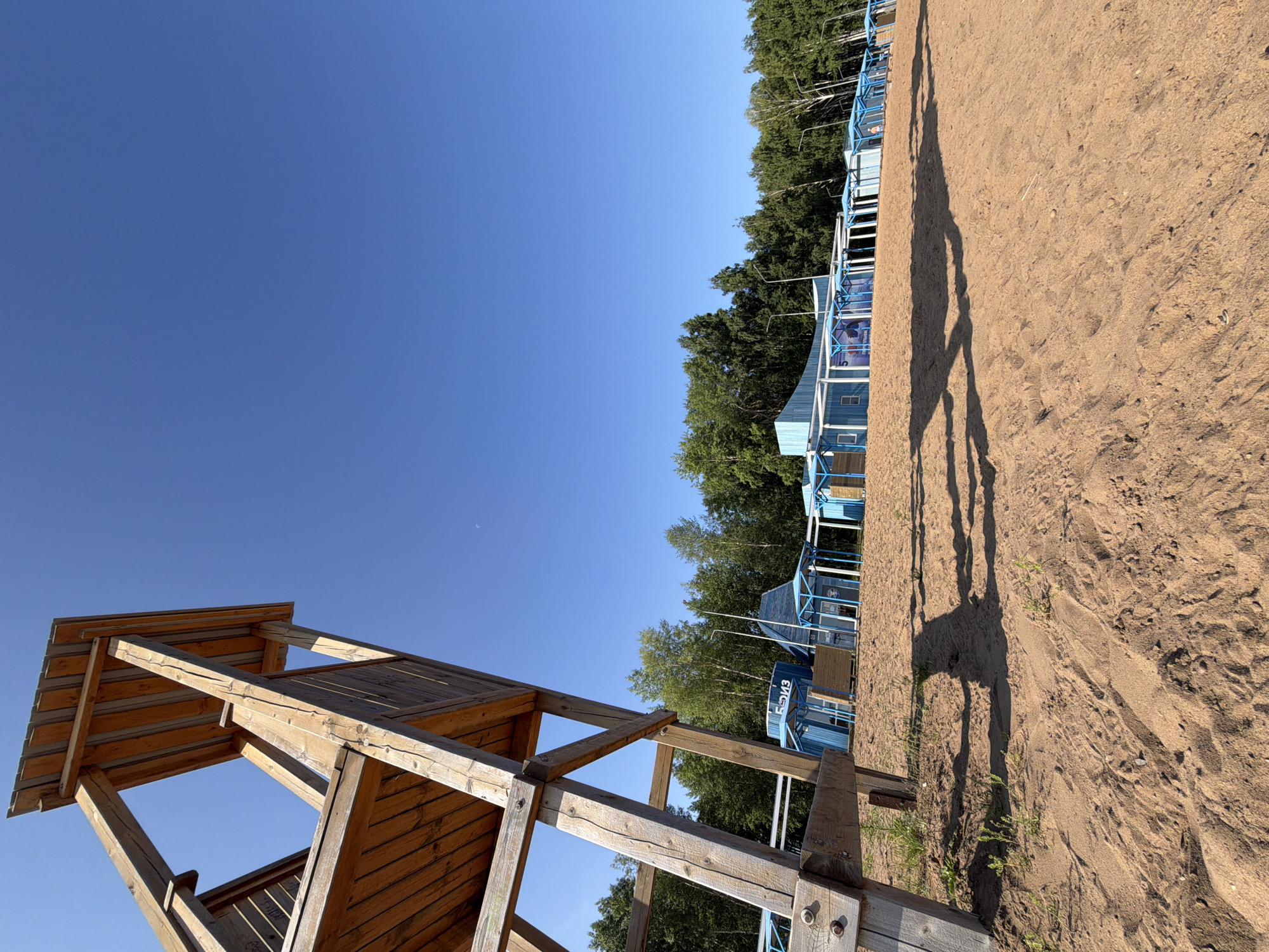 A low-angle, wide-shot photograph captures a sandy beach area with a wooden lifeguard tower in the foreground, leaning towards the left of the frame. The tower is constructed from rough-hewn wooden planks and beams. Behind the tower, a row of small, light blue, open-sided structures with blue awnings stretches horizontally across the mid-ground. These structures appear to be changing rooms or beach cabanas, some displaying signage in Russian. A line of trees forms a dense green backdrop behind the blue structures. The sky is a clear, bright blue with a faint crescent moon visible. The sandy ground in the foreground casts long shadows from the lifeguard tower, indicating a bright sunny day. The scene is likely part of a beach resort or recreational area, possibly located near the Kama River in Chaikovsky, Russia, a city known for its water-based recreational facilities. There are no visible people in the immediate foreground, but a few figures can be seen in the distance near the blue structures.