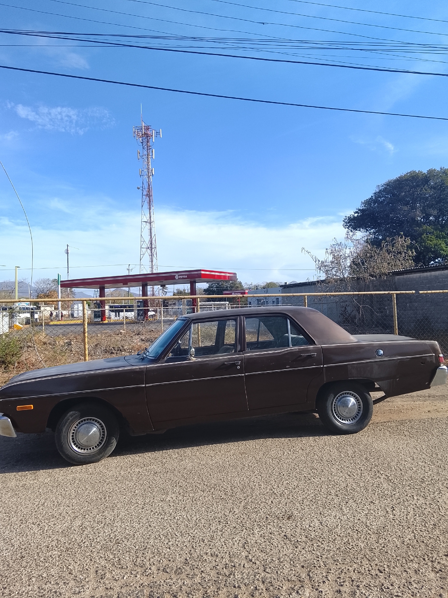 The image shows a brown, vintage four-door sedan parked on a paved road. Behind the car is a wire fence. Behind the fence, there's a gas station with a red and white awning, the logo of which is indistinct but potentially PVDSA. A cell tower is visible behind the gas station. The location appears to be Altagracia, Venezuela. The sky is blue with scattered clouds, and the overall lighting suggests it is daytime.