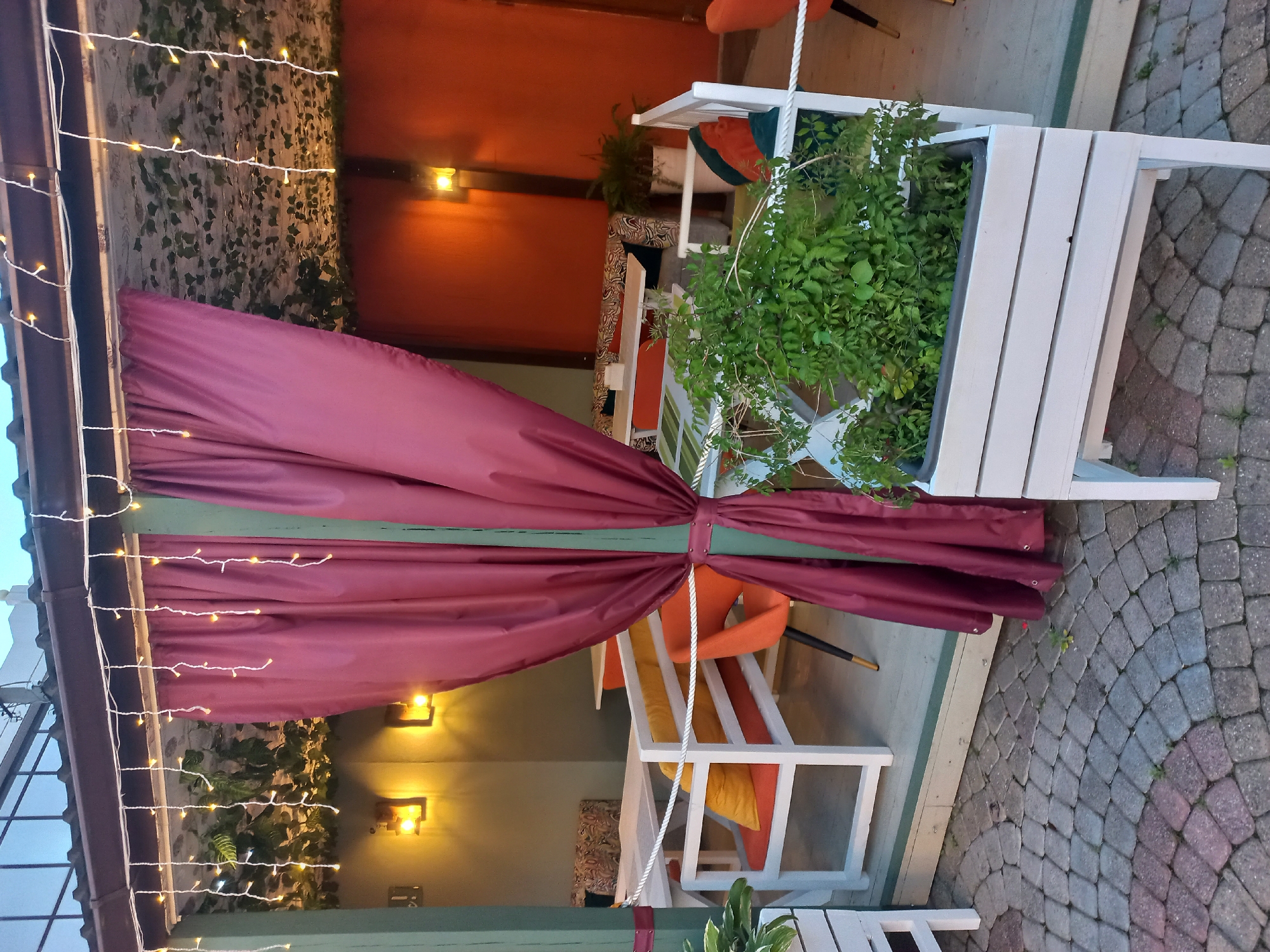 The image depicts a patio in Sochi, Russia. A maroon-colored curtain is tied back, revealing outdoor seating with orange and yellow cushions. String lights illuminate the space, which is decorated with greenery. The patio is paved with gray cobblestones.
