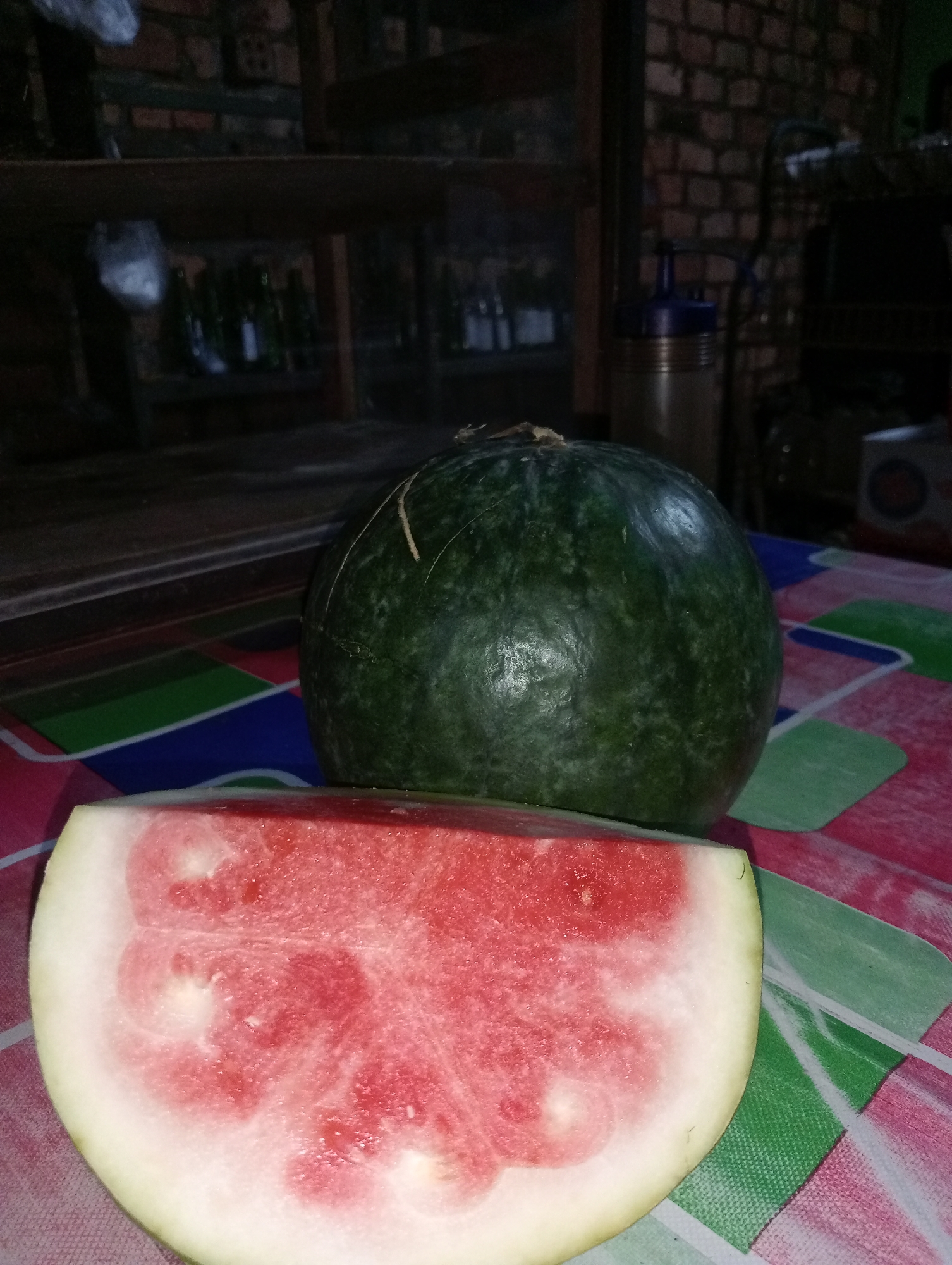 An outdoor scene at dusk features a ripe watermelon on a patterned tablecloth. The watermelon is dark green with light green stripes. A slice of watermelon, revealing its pink flesh and light-colored rind, is positioned in the foreground. In the background, a dark wooden shelf holds several bottles, suggesting a storage area or possibly a kitchen. The lighting is dim, indicating it's likely evening. The environment appears to be a residential area in Kota Negara Timur, Indonesia.