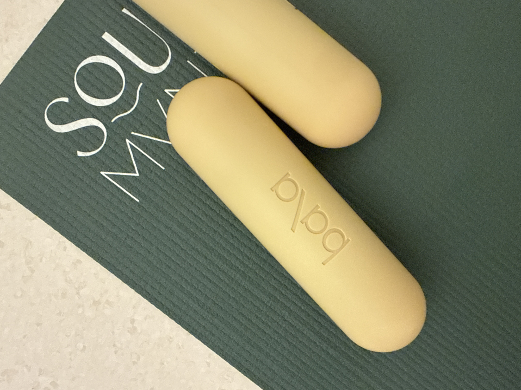 The image shows two pale yellow Bala Bangles resting on a green and white yoga mat. One bangle is prominently in the foreground, displaying the "bala" brand name in recessed lettering. The second bangle is partially visible in the upper right corner. The yoga mat has white lettering "SOU MY" partially visible on the left side. The mat rests on a light beige surface. The setting appears to be indoors with soft, diffused lighting. The scene does not provide sufficient information to determine its location in Dubai or any activity taking place.