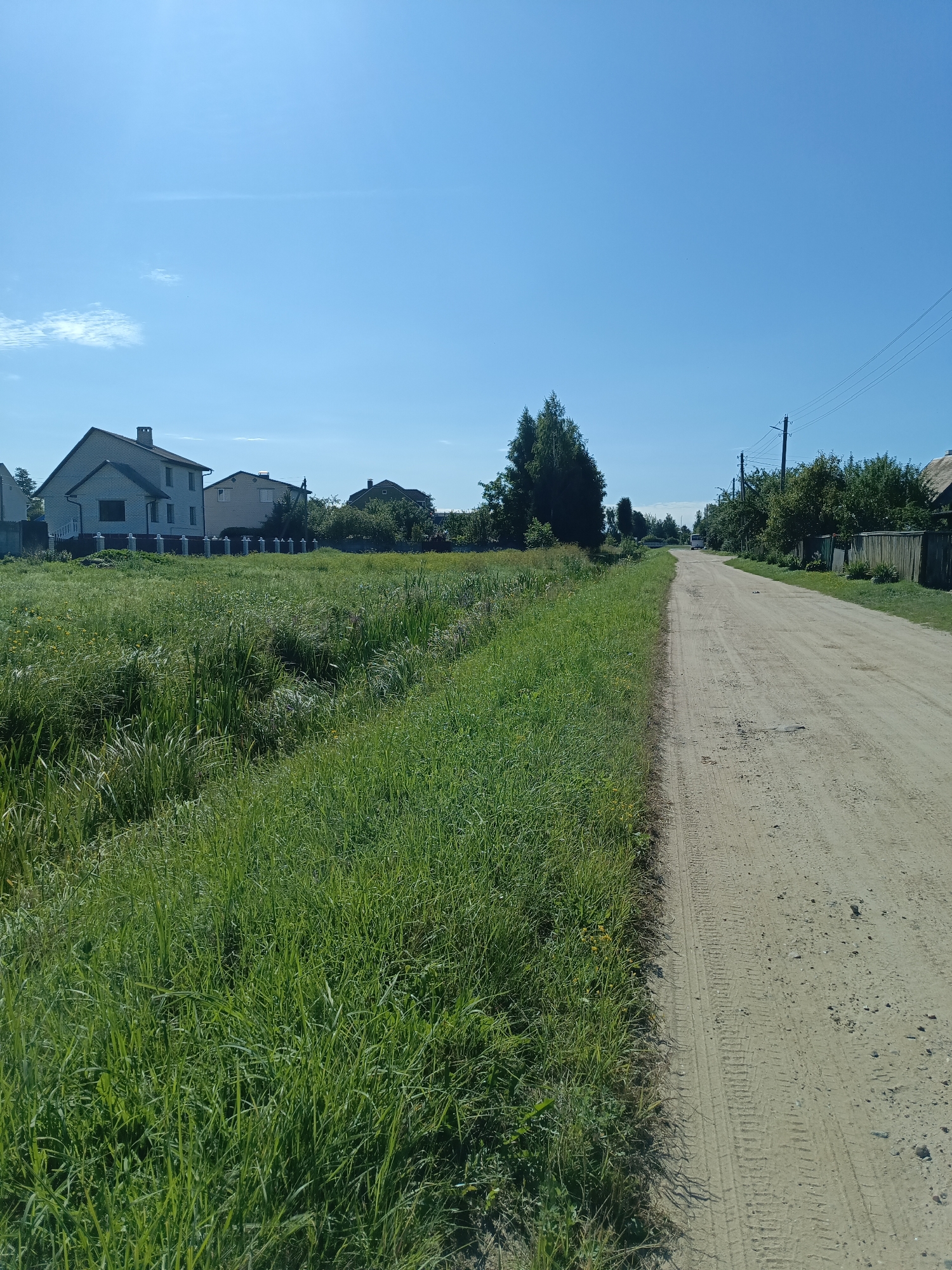 The image depicts a rural scene on a bright, sunny day. A dirt road runs from the foreground to the background, curving slightly to the right. On the left side of the road, there is a dense field of tall green grass with some yellow flowers. Beyond the field, several houses with tiled roofs are visible. The houses appear to be residential properties, with one larger brick house in the left mid-ground. In the distance, more trees and houses can be seen, suggesting a village or a suburban area. Utility poles with wires line the right side of the road, indicating the presence of electricity infrastructure. The sky is clear and blue with a few faint wisps of clouds. The overall impression is one of a tranquil, rural environment. The location is identified as being in the city of Ivatsevichy, Belarus.