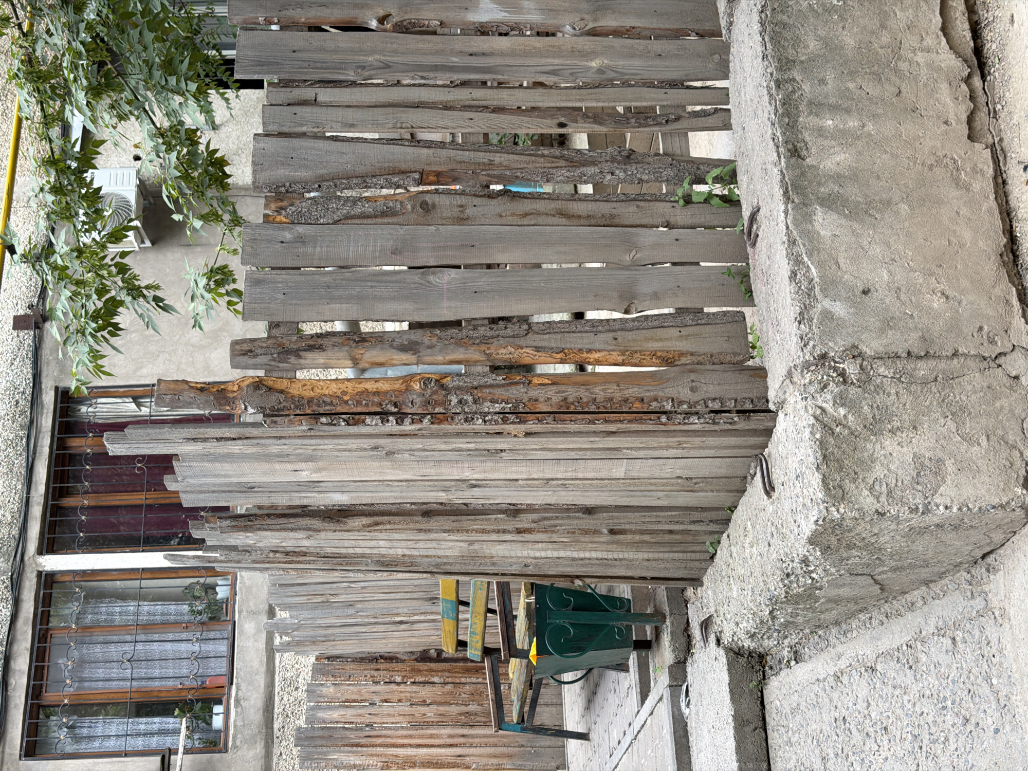 The image depicts an outdoor scene with a rustic wooden fence in the foreground. The fence is constructed from irregularly shaped, weathered wooden planks of various widths and heights, some with bark still attached. Behind the fence, to the left, is a two-story building with two windows on the upper level. These windows have metal security bars and white lace curtains. The building's facade is a textured concrete or stucco. To the right of the fence, a tree with green leaves extends into the frame, and an air conditioning unit is visible on the building's wall.
In the middle ground, behind a section of the fence, a simple park bench with a yellow and blue seat is visible. Next to the bench is a dark green metal trash can. The ground in front of the bench appears to be paved with rectangular stones.
The foreground features a large, rough, concrete block. This block has a textured, weathered surface and exposed aggregate. Two metal loops are embedded in the top surface of the concrete block, facing outwards. The ground around the concrete block is made of rough concrete or asphalt. The overall impression is of an urban or semi-urban environment, possibly an alleyway or a courtyard, with elements of disrepair or a DIY aesthetic.
This scene is located in the city of Shymkent, Kazakhstan.