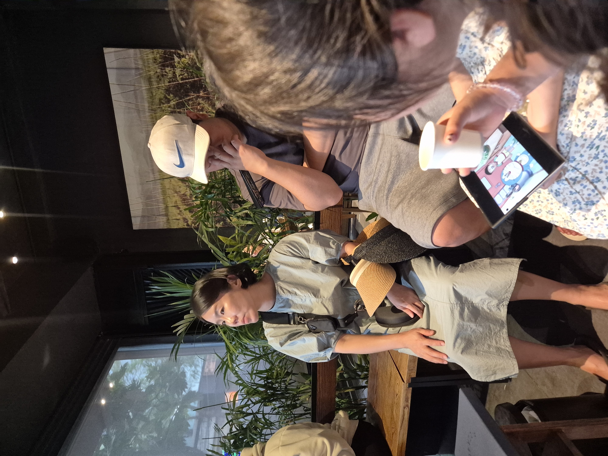 The image depicts an indoor scene, possibly a cafe, with three individuals. On the left, a woman in a light blue dress is seated, looking to her left. She is carrying a straw hat and a black purse. In the center, a man wearing a white Nike cap and a grey t-shirt is seated, with his hand near his face. To the right, a person in a floral dress is looking down at a smartphone, holding a white cup. The background shows plants and a large framed artwork. The lighting suggests it is daytime. The presence of plants and the general ambiance suggest a relaxed, possibly a coffee shop or restaurant setting. There is no indication of the specific location in Da Nang, Vietnam.
