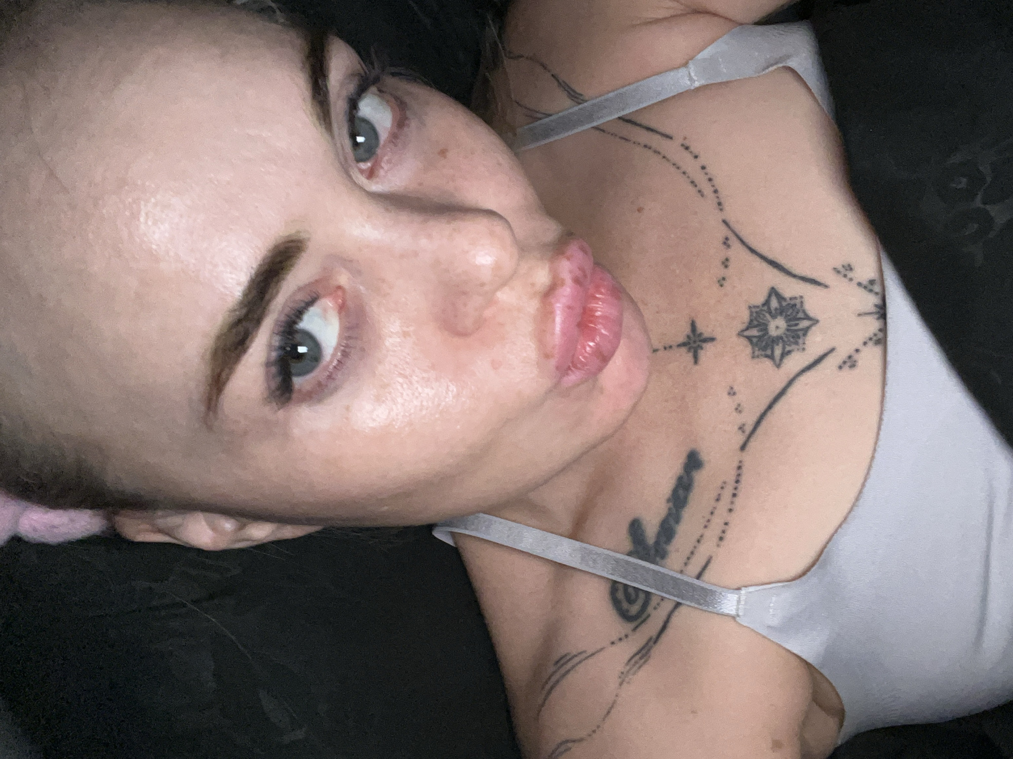 A close-up, high-angle, slightly over-the-shoulder selfie shows a woman with fair skin, blue-green eyes, and full lips. She is wearing a light gray bra with thin straps. Her hair is pulled back with a pink accessory visible at the top of her head. She has a tattoo on her upper chest, a line of text on her left collarbone and a starburst design with radiating lines and dots centered on her sternum. The background is dark, suggesting an indoor or nighttime setting. There is no discernible city or location information present in the image.