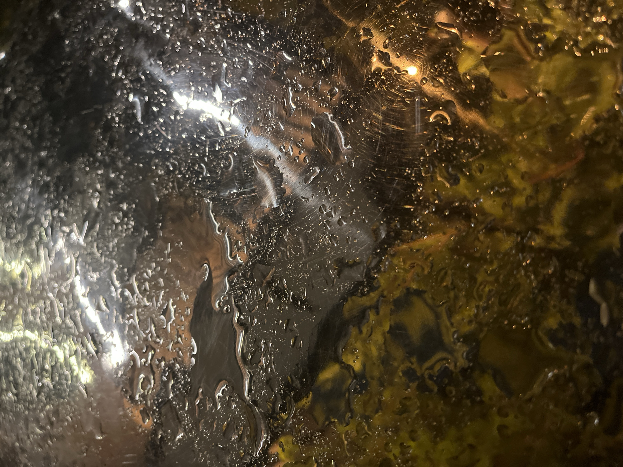 The image depicts a wet glass surface, likely a window, with raindrops and water streaks obscuring the view. Reflected lights and shapes are visible through the glass, suggesting an urban environment at night. The reflections are predominantly yellow and dark, perhaps indicating streetlights and foliage. The scene evokes a rainy night in a city. There is no evidence to suggest that this image depicts a location in Krasnoyarsk, Russia.