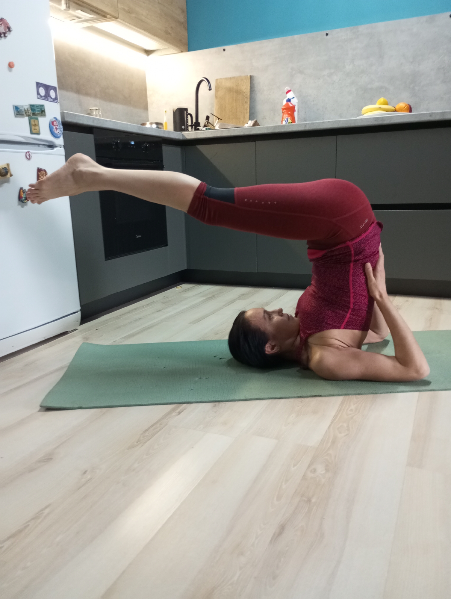 A woman is performing the Halasana yoga pose, known as plow pose, on a green yoga mat inside a kitchen. The woman is wearing a maroon-colored exercise top and leggings. She is lying on her back with her legs raised and extended over her head, her feet visible near the refrigerator. Her hands are supporting her back. The kitchen has light-colored wood-look flooring. The cabinets are dark gray with a light gray countertop. There's a sink with a black faucet, and some household items and fruit are on the counter. A white refrigerator is visible on the left with magnets on it. The location is unknown, so I can not confirm the location to be in the city of Pervomaysky, Russia.