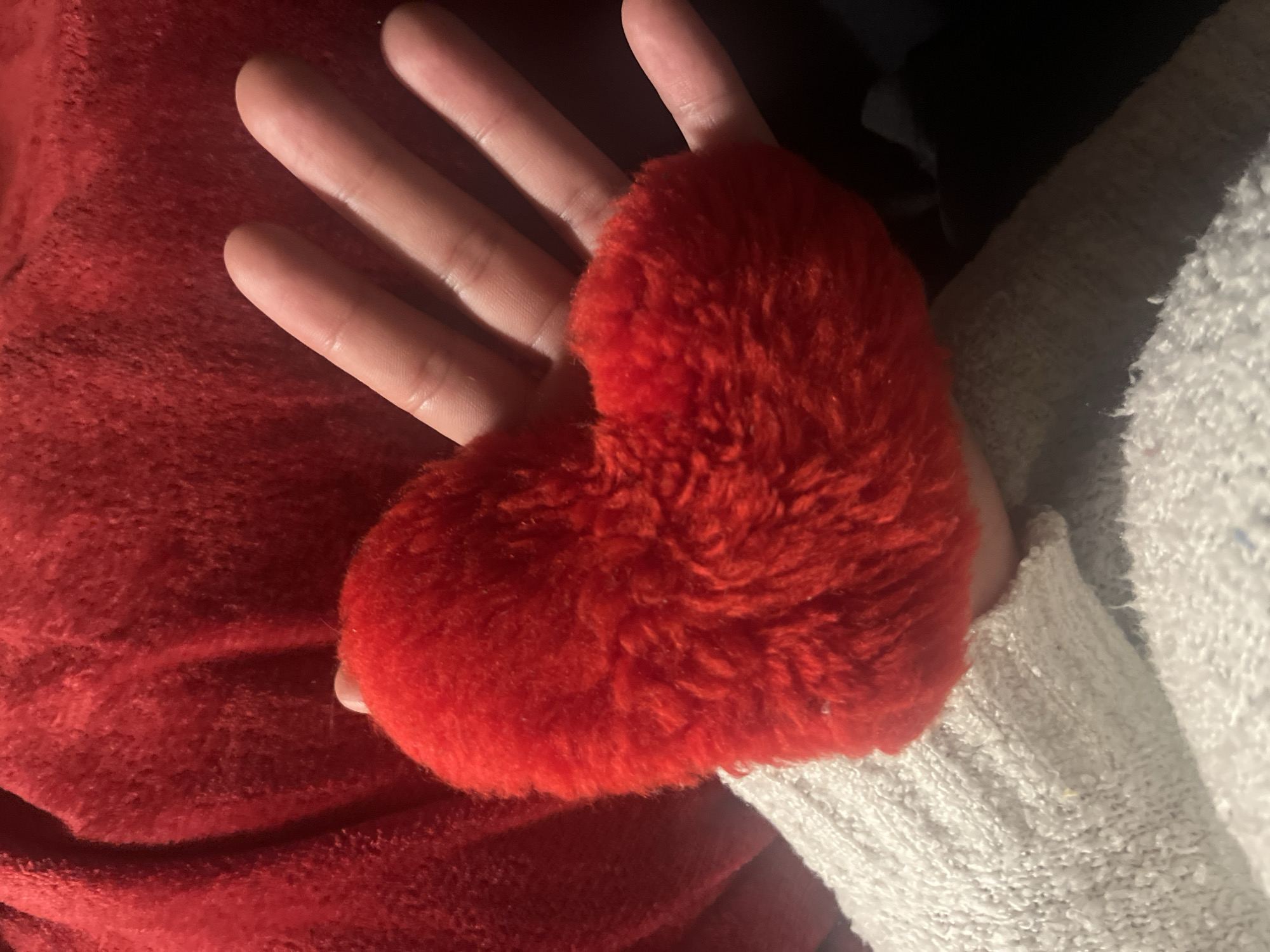 A hand holds a red, fluffy heart-shaped object. The hand has pale skin and is resting on a red fuzzy blanket. A white, knitted sleeve is partially visible next to the hand and blanket.