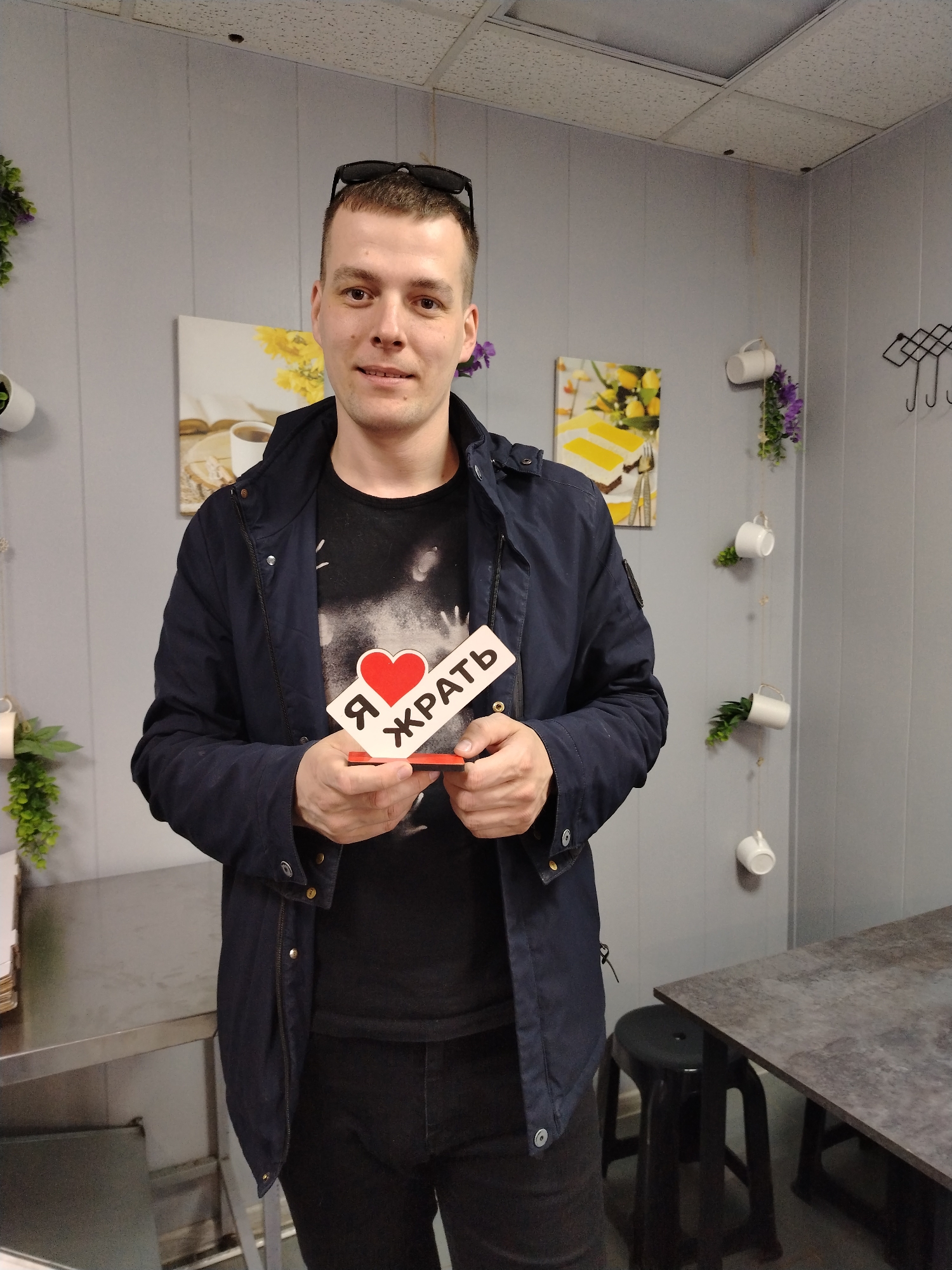 A man stands indoors, holding a sign that says "Я ❤️ ЖРАТЬ" (I love to eat). He is wearing a dark blue jacket, a black t-shirt, and black jeans. He has short brown hair and sunglasses on his head. The background includes gray walls with decorative elements such as paintings, hanging cups with plants, and hooks. There is a table and stool visible on the right. The image appears to be taken in the city of Chaikovsky, Russia, based on the Russian text on the sign.