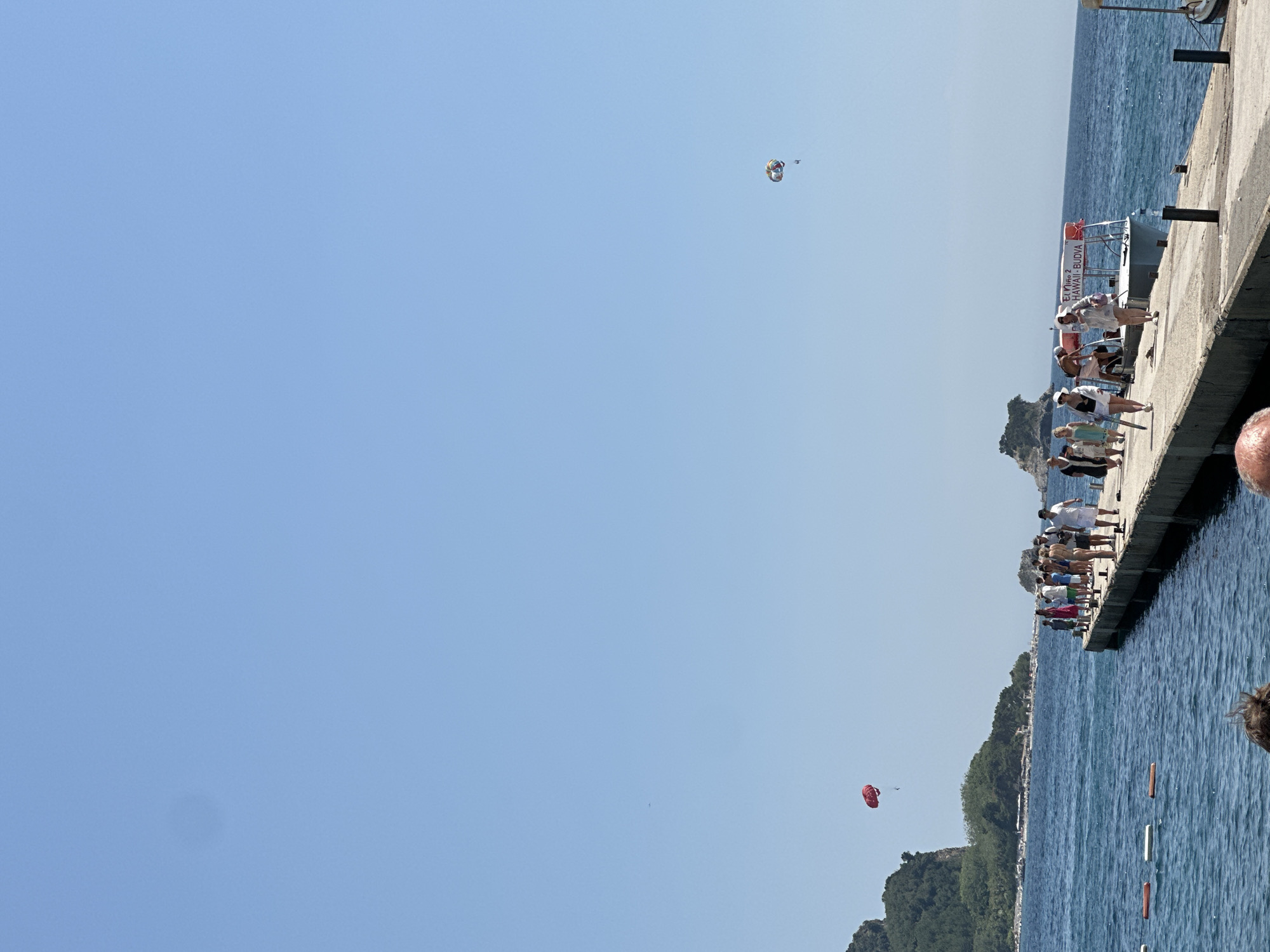 The image depicts a sunny day at a coastal location, likely in Budva, Montenegro, given the text on the boat. Several people are gathered on a concrete pier, which extends into the blue water. The pier is lined with boats, one of which has the text "AMAZING BEACH" and "HAWAII - BUDVA" visible on its side. Some individuals are standing, possibly waiting for or observing activities, while others are seated or appear to be involved in some preparations.
In the sky, there are at least two parasails, suggesting parasailing is taking place as a recreational activity. One parasail, a multi-colored canopy, is higher in the sky, while another, a bright red one, is closer to the water and a tree-covered hillside. The water is calm with gentle ripples, reflecting the clear blue sky. The presence of lush green vegetation on the hillsides indicates a warm, pleasant climate. The scene conveys a sense of vacation and leisure, with people enjoying outdoor water sports and the seaside atmosphere. The time of day appears to be midday, given the bright sunlight and clear skies.