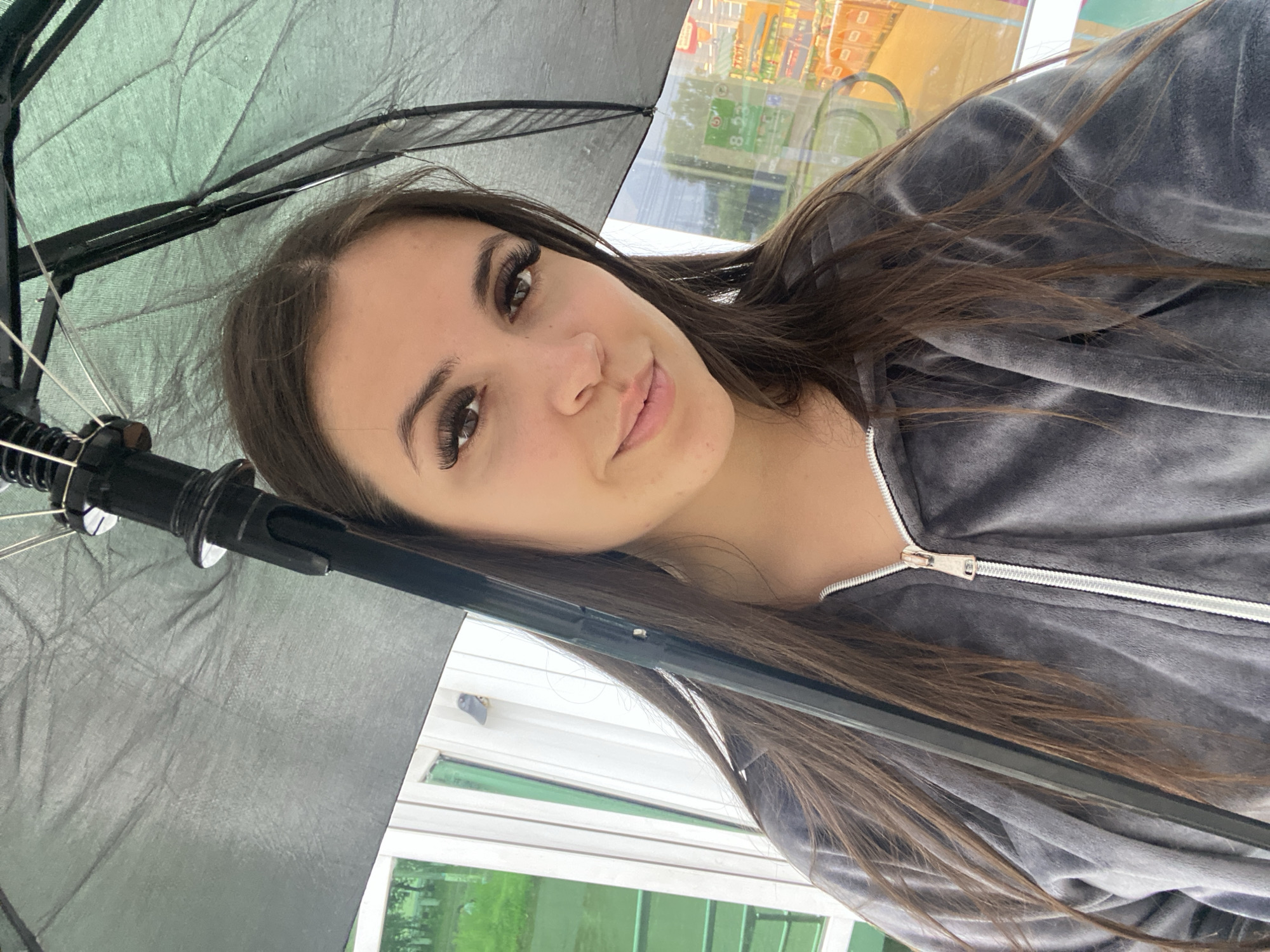 A woman is taking a selfie outdoors, holding a black umbrella over her head. She is wearing a gray velour zip-up hoodie. The background shows a building with large windows and some greenery. The lighting suggests it might be a cloudy or overcast day. Based on the surrounding context of the image, the location is believed to be in the city of Chaikovsky, Russia.