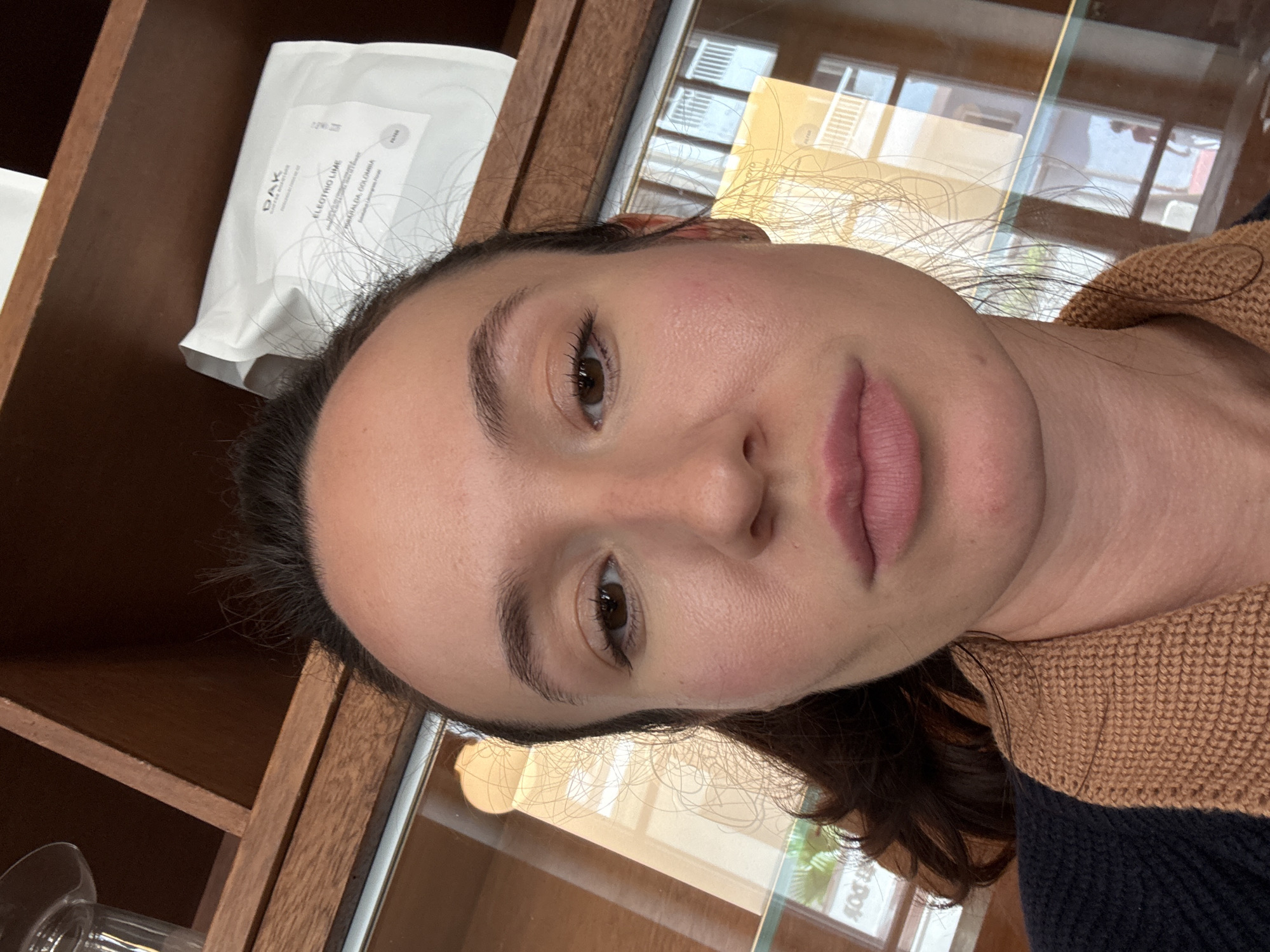 A selfie depicts a woman with brown hair and eyes, and light skin. Her facial features are prominent, including defined eyebrows and lips. She's wearing a brown knitted collar over a navy blue top. In the background, a wooden shelving unit is partially visible, containing a white package labelled "ELECTRIC LIME" and "SANTA MARTA, COLOMBIA," along with a glass object. A window reflects what seems to be an urban environment with other buildings and greenery, possibly suggesting a location within Tel Aviv, Israel.