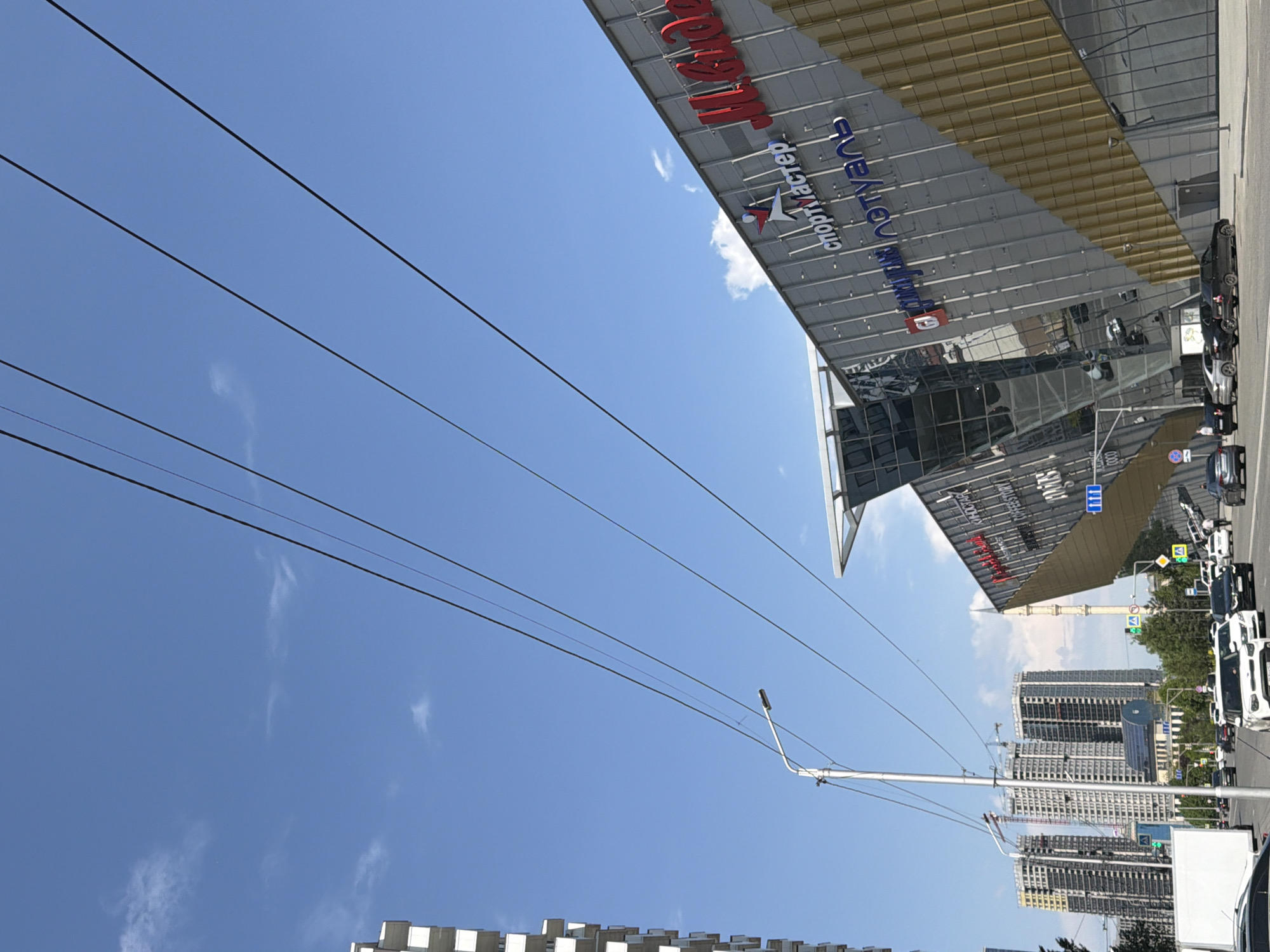 The image shows a street view in Grozny, Russia, with the "Sportmaster" store visible. The sky is blue with some clouds. Power lines run across the sky. The street is lined with buildings and cars. There is a streetlight on the side of the road. Tall apartment buildings are visible in the background. A traffic light is visible on a traffic pole.