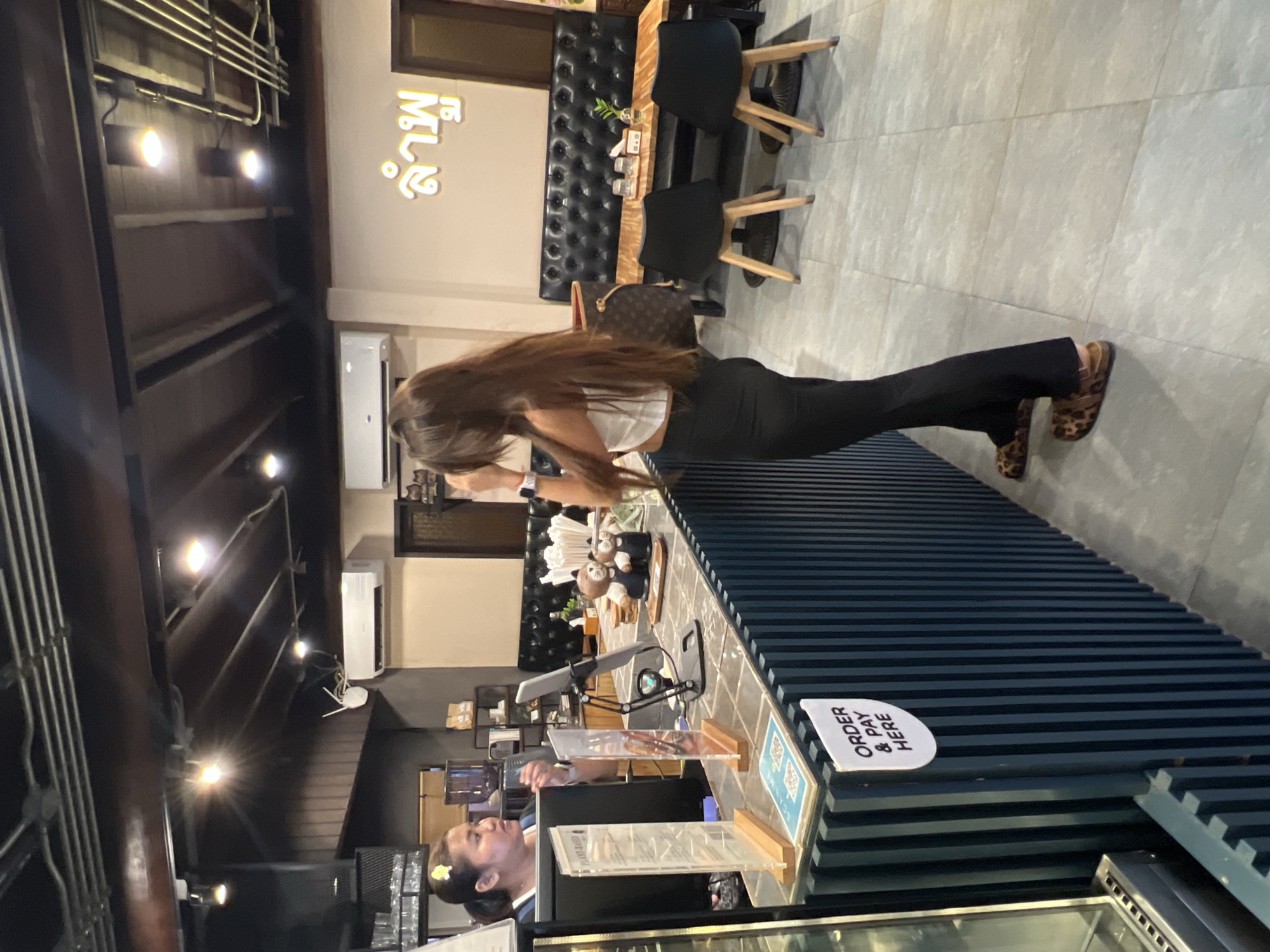 A woman with long brown hair is standing on her hands, in a plank position, on a counter in what appears to be a restaurant or cafe. She is wearing black pants and a white top. She is in the middle of the frame, her body parallel to the floor. Her legs are extended behind her. She is wearing leopard-print shoes.
Behind her, the counter has various items, including what looks like a display of snacks or drinks, and some small decorative figures. There are also menus in clear plastic holders on the counter. To the left of the woman, a person is visible behind the counter, possibly an employee, wearing a yellow hair clip. They are looking towards the woman.
The setting appears to be an indoor establishment with a modern design. The walls are a light color, and there is a prominent neon sign with Thai script that reads "กินละมุน" (Gin Lamun). The floor is tiled with large, light-colored tiles. The furniture includes dark leather banquettes and wooden chairs with black seats. The lighting is warm, suggesting it could be evening or a dimly lit interior. A sign on the counter reads "ORDER & PAY HERE." The overall impression is a casual dining or service establishment.