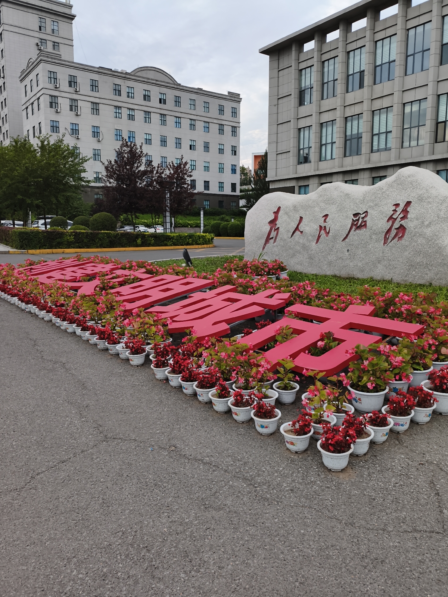 In the Jiguan District of China, a large, grey, carved stone rests behind a prominent red inscription. The inscription consists of large, three-dimensional red characters spelling out "为人民服务," which translates to "Serving the People." These characters are arranged in a line and are further embellished with numerous potted red and pink begonia flowers in white planters, creating a vibrant floral border.
To the left of the stone and inscription, a modern, light-colored building with multiple windows dominates the background. This building appears to be institutional or governmental in nature, with a taller section extending upwards. To the right, another building of similar architectural style is visible, also featuring numerous windows. The environment suggests an outdoor public space, possibly in front of a government office or university. The ground is paved with asphalt, showing some minor cracks. The sky is overcast, indicating a cloudy day. The overall scene conveys a sense of civic pride and public service.