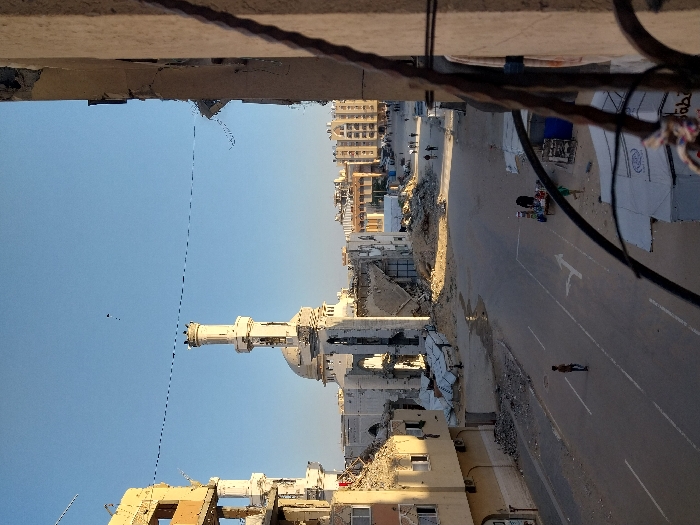 The image shows a street scene from an elevated perspective. Buildings line both sides of the street, with significant damage and debris visible. A prominent white mosque with a tall minaret stands out amidst the rubble. A few people are seen walking or standing on the road. Overhead cables and part of a damaged building frame the top of the picture, suggesting the viewpoint is from a nearby structure. The sky is clear and blue.