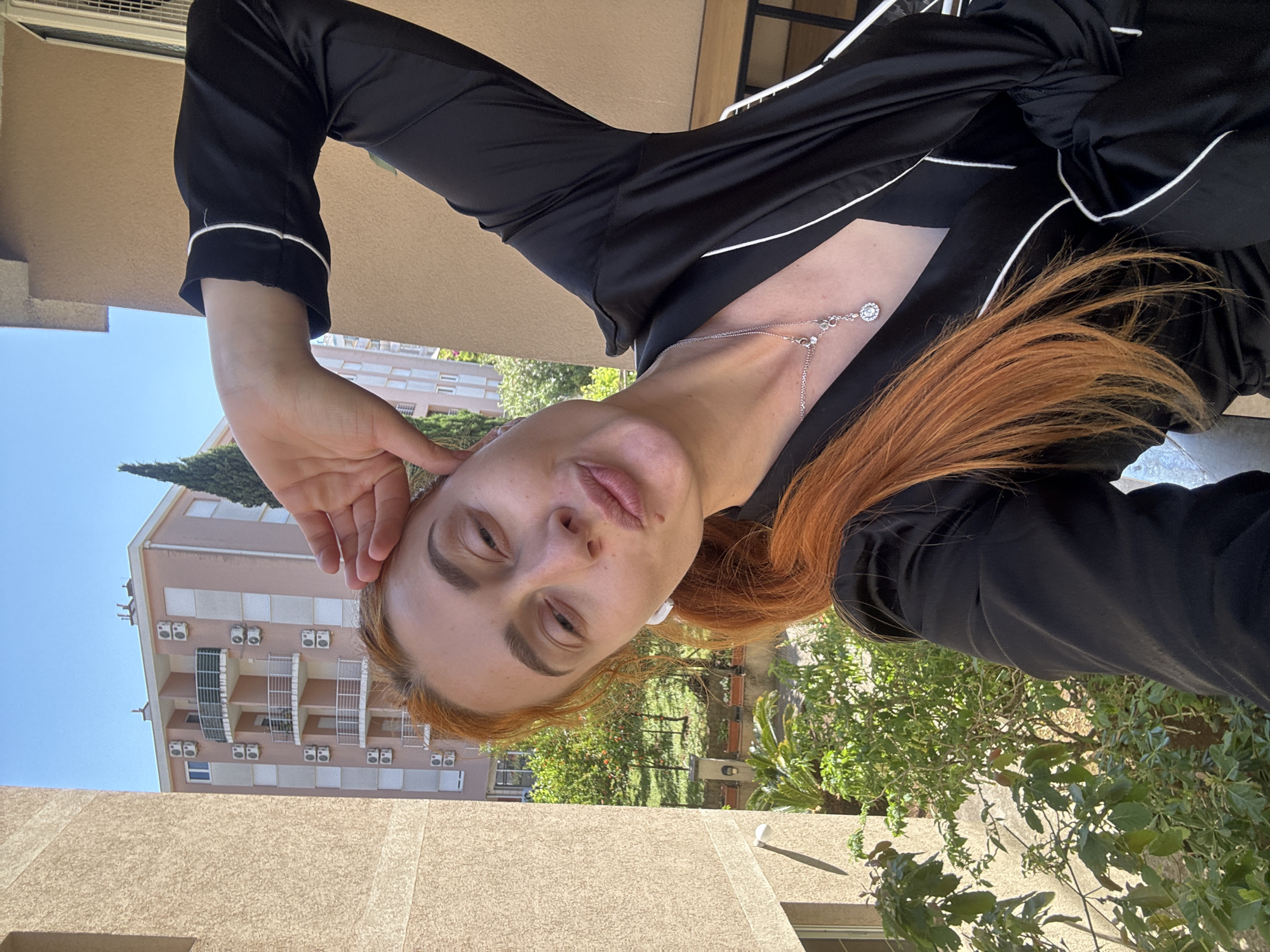 A young woman with fair skin and reddish-blonde hair is looking at the camera with a direct gaze. She is wearing a black silky pajama-style top with white piping. A delicate silver necklace adorns her neck. Her left hand is raised near her head.
Behind her, the environment reveals a sunny urban landscape. A multi-story apartment building with tan-colored walls and rows of air conditioning units on each level is visible. Green trees and bushes are also present, suggesting a residential area.
Based on the provided context and the architectural style of the building, the location is most likely within the city of Budva, Montenegro.