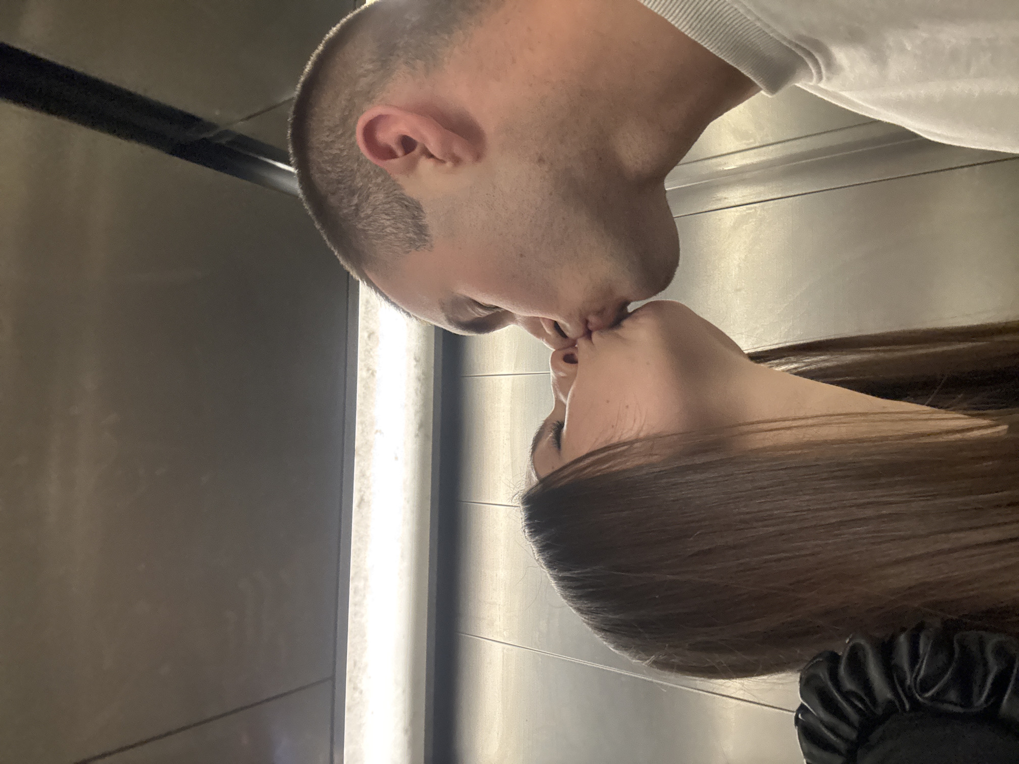 The image shows two individuals, a man and a woman, engaged in a kiss. The photo is taken from a close-up, side profile perspective, making their faces prominent. The man, positioned on the left, has very short, shaved hair and a visible ear. He is wearing a light-colored, possibly white, long-sleeved shirt or sweatshirt, part of which is visible on the upper right. The woman, on the right, has long, dark brown hair that cascades down her back. A black scrunchie or similar hair accessory with a textured, possibly satin or leather-like, finish is visible near her shoulder or neck area. Both individuals have their eyes closed as they kiss.
The setting appears to be an indoor space with plain, possibly metallic or dark paneled walls, creating a somewhat enclosed environment, possibly an elevator or a modern hallway. There are horizontal lines suggesting paneling or reflections on the walls. The lighting is artificial and seems to come from above or the side, creating some highlights on the surfaces. The overall mood conveyed is one of intimacy and affection. There is no discernible text visible in the image.