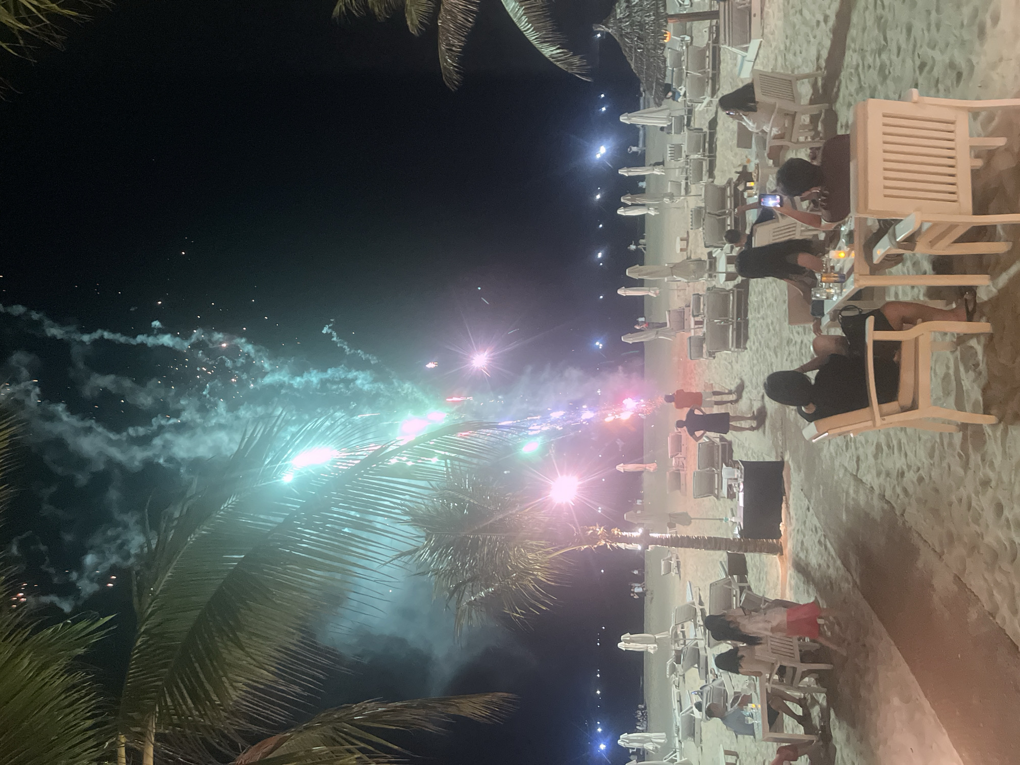 The image captures a nighttime scene on a beach in Da Nang, Vietnam, with fireworks illuminating the sky. In the foreground, people are seated at tables on the sand, some appearing to dine. Palm trees frame the scene, their fronds silhouetted against the dark sky. Beyond the immediate seating area, rows of empty lounge chairs and umbrellas suggest a resort or public beach. In the distance, the coastline is dotted with lights, indicating the presence of buildings or other structures. The fireworks display is the dominant visual element, with streaks of green, pink, and blue light exploding across the sky, casting an ethereal glow on the surroundings and the smoke trailing from the bursts. The overall atmosphere is one of celebration and leisure, set against the backdrop of a tropical night.