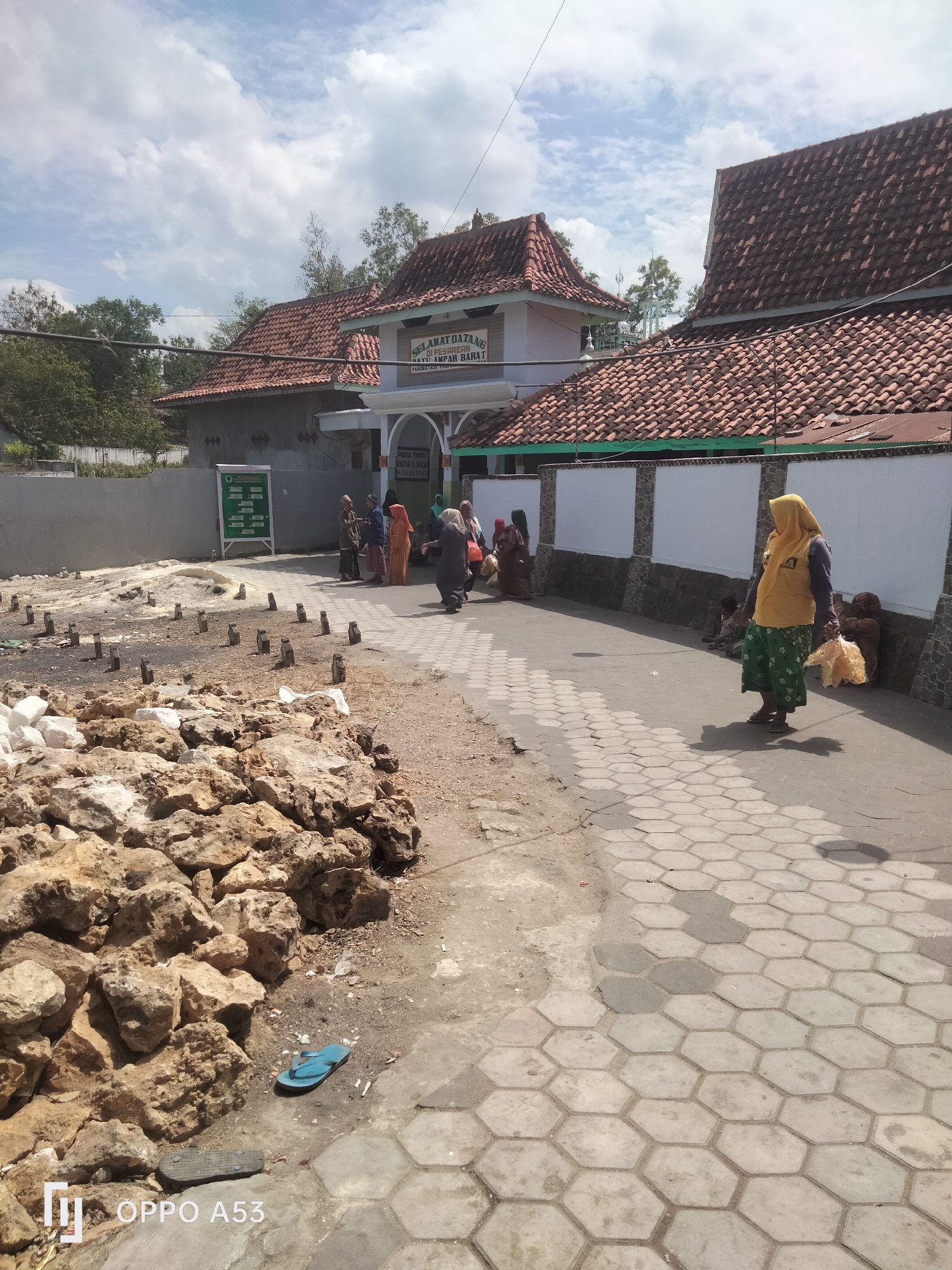 This is an outdoor, eye-level, full-shot photograph taken on a sunny day. The image depicts an entrance area with a tiled walkway leading towards buildings with red tile roofs. To the left, there is a pile of large rocks and what appears to be a graveyard with small, square markers. Several people are present, including a woman in a yellow hijab and green skirt walking towards the right side of the frame, carrying a bag. Other individuals, some wearing traditional Islamic clothing, are standing or sitting near the buildings. A sign with text in Indonesian is visible above an arched entrance. The environment appears to be dry and dusty. The location is the Badung Cemetery in Badung, Indonesia.