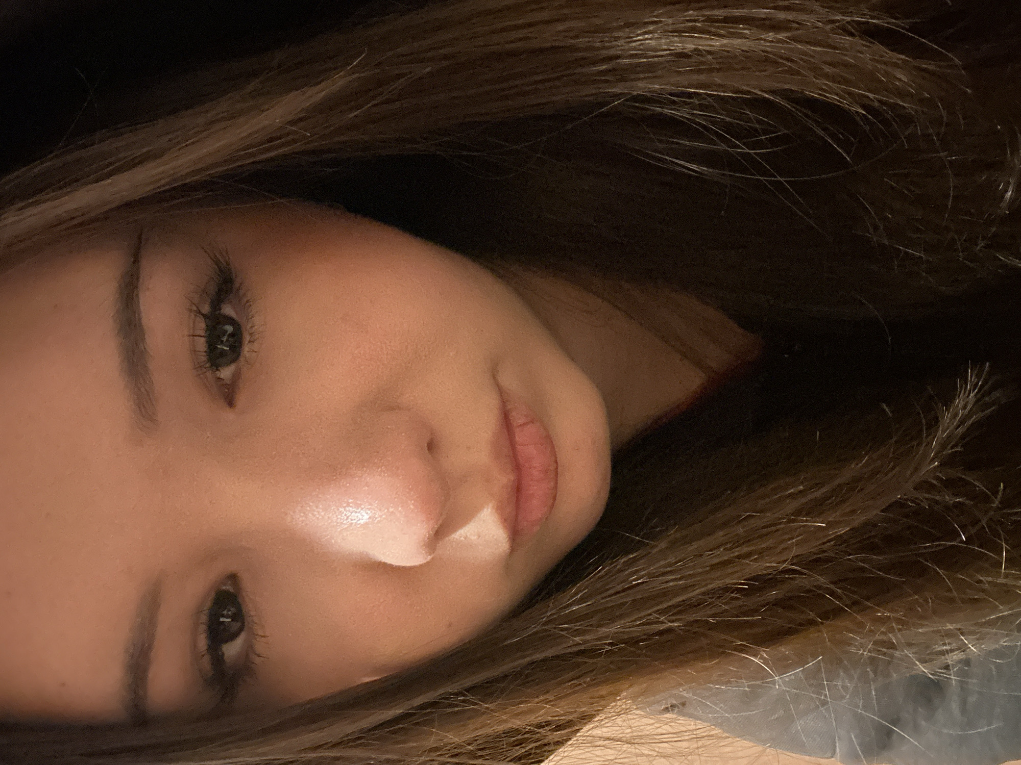 A close-up, head-and-shoulders portrait of a young person, likely female, captured in the city of Naberezhnye Chelny, Russia. The subject possesses a fair to medium complexion and long, straight to wavy brown hair with lighter, possibly blonde, highlights, framing the face and extending over the shoulders. Both eyes are visible, directed downwards or slightly forward, with prominent, dark eyelashes. Well-defined eyebrows are present above the eyes. The nose is centered, exhibiting a highlight along its bridge. The lips are closed in a slight pout, displaying natural texture. The overall facial expression suggests a solemn or contemplative state. The lighting is warm and directional, illuminating the forehead, bridge of the nose, and lips, creating highlights on the skin and on individual hair strands. The background is dark and out of focus, offering no discernible environmental features. The subject is wearing a dark garment, possibly a hoodie, with a portion of lighter grey fabric visible on the lower left side of the frame.