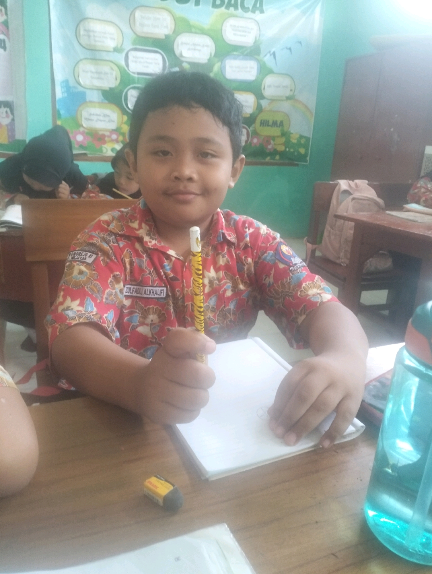 The image depicts a young boy sitting at a wooden desk in what appears to be a classroom or a learning environment. The boy is wearing a red shirt with a repeating floral pattern and a name tag that reads "ZULFADLI ALKHALIFI". He is holding a yellow pencil with a tiger-like pattern and appears to be drawing or writing on a white piece of paper. An eraser is visible on the desk in front of him. In the background, there are other students, some partially obscured, and a banner with text and drawings. To the right of the boy, a blue water bottle with a straw is present. The overall setting suggests a school in Blabakan, Indonesia, given the context provided.