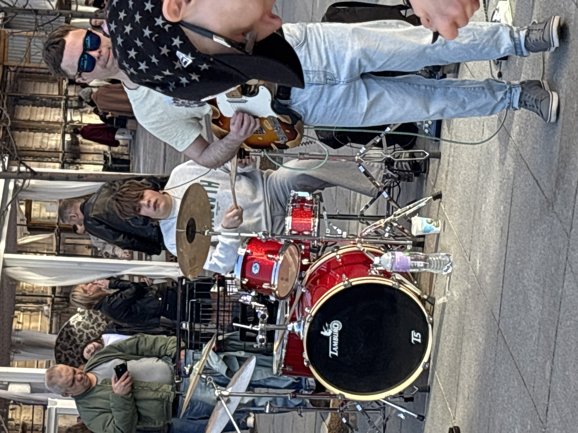 A street band performs outdoors on a gray paved surface, possibly in Saint Petersburg, Russia, given the architecture and people present. A drummer, wearing a light gray sweatshirt, sits behind a red drum set, holding drumsticks. A guitarist wearing a white shirt, light blue jeans, and gray sneakers is partly visible from the waist up. He also wears a black hat with white stars and sunglasses. A microphone is near his mouth. Around the musicians are several observers, including a man in a green jacket using a smartphone, and others who are blurred. White fabric is seen above the group and observers, likely part of some sort of structure that is open to the elements.
