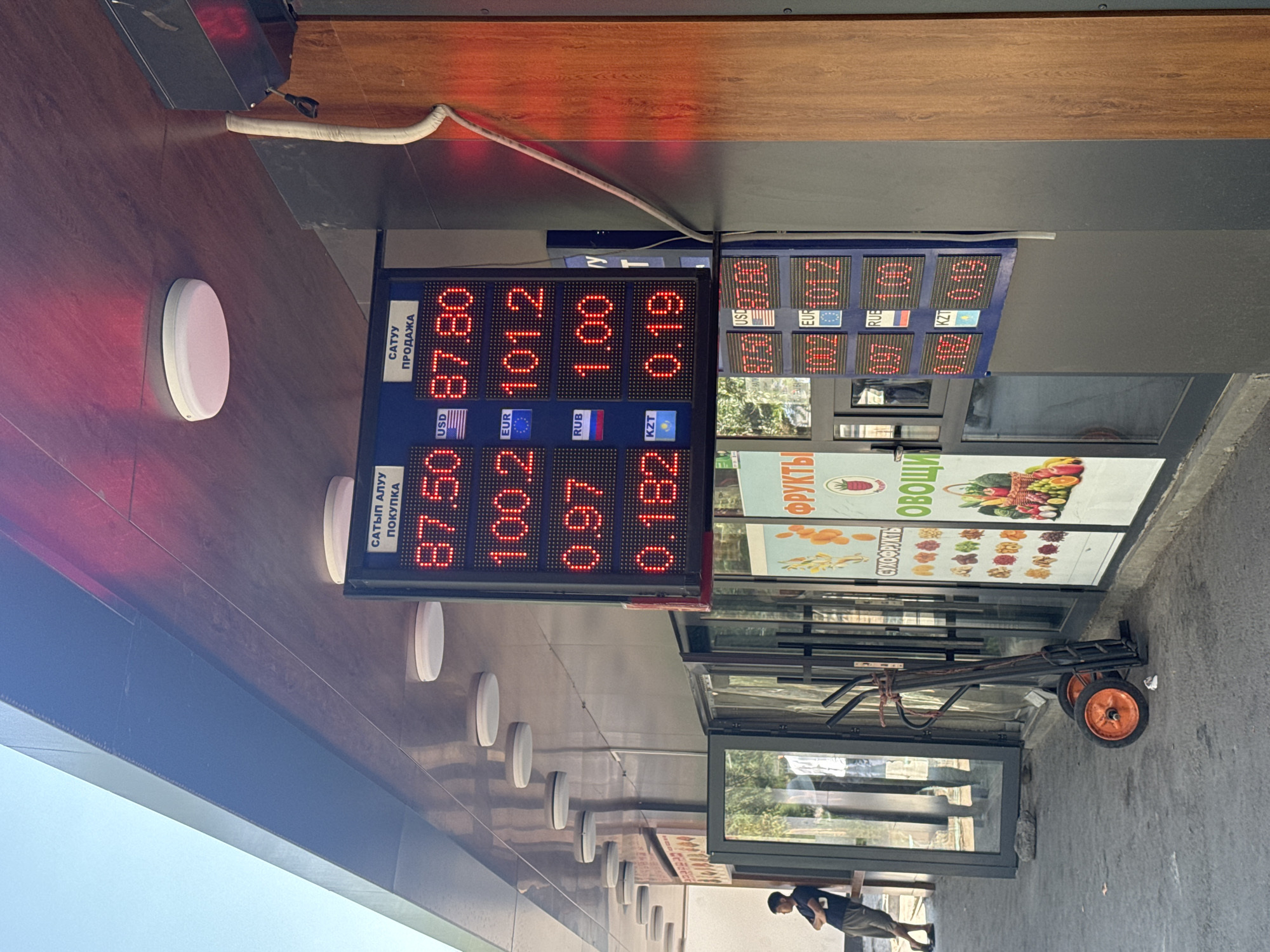 The image shows a digital currency exchange rate board displayed on the exterior of a building, likely in Bishkek, Kyrgyzstan. The board is blue and features red LED digits displaying exchange rates for USD, EUR, RUB, and KZT. The top row indicates "SATIP ALUU" (BUY) and "SATUU" (SELL). For USD, the buy rate is 87.50 and the sell rate is 87.80. For EUR, the buy rate is 100.2 and the sell rate is 101.2. For RUB, the buy rate is 0.97 and the sell rate is 1.00. For KZT, the buy rate is 0.182 and the sell rate is 0.19. Below the currency exchange board, there is a colorful advertisement for fruits and vegetables. To the left of the building, a young man wearing a dark t-shirt and shorts is leaning against a wall and looking towards the right. A dark-colored hand truck with orange wheels is parked on the pavement in front of the building. The environment appears to be an outdoor commercial area with a paved walkway and glass storefronts. The lighting suggests it is daytime.