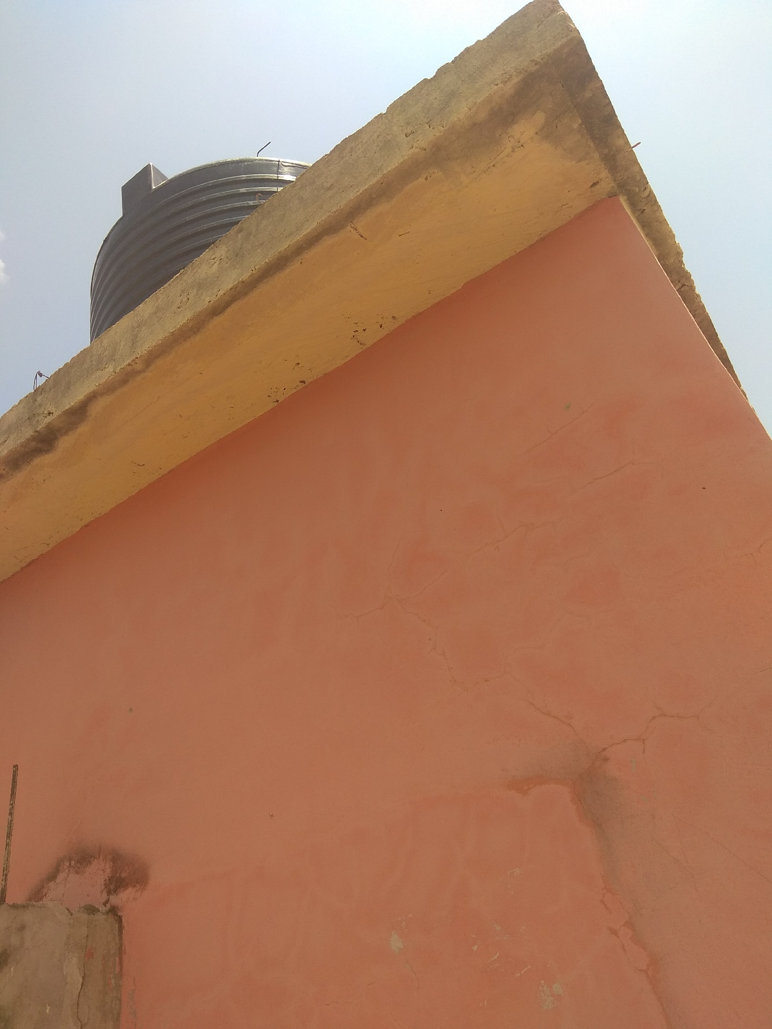 The image shows a close-up view of the exterior of a building with a water tank on the roof. The building has a peach-colored wall that shows some signs of age and weathering, with visible cracks and water stains. A concrete ledge or overhang runs along the top of the wall, painted yellow. A black plastic water tank is visible on the roof behind the ledge, partially obscured. The sky is clear and bright, suggesting it is daytime, likely a sunny day. The perspective is from below, looking upwards towards the roof and sky. There are no people visible in the image. The scene is likely residential or commercial, common in Jalingo, Nigeria, where such water storage systems are prevalent.