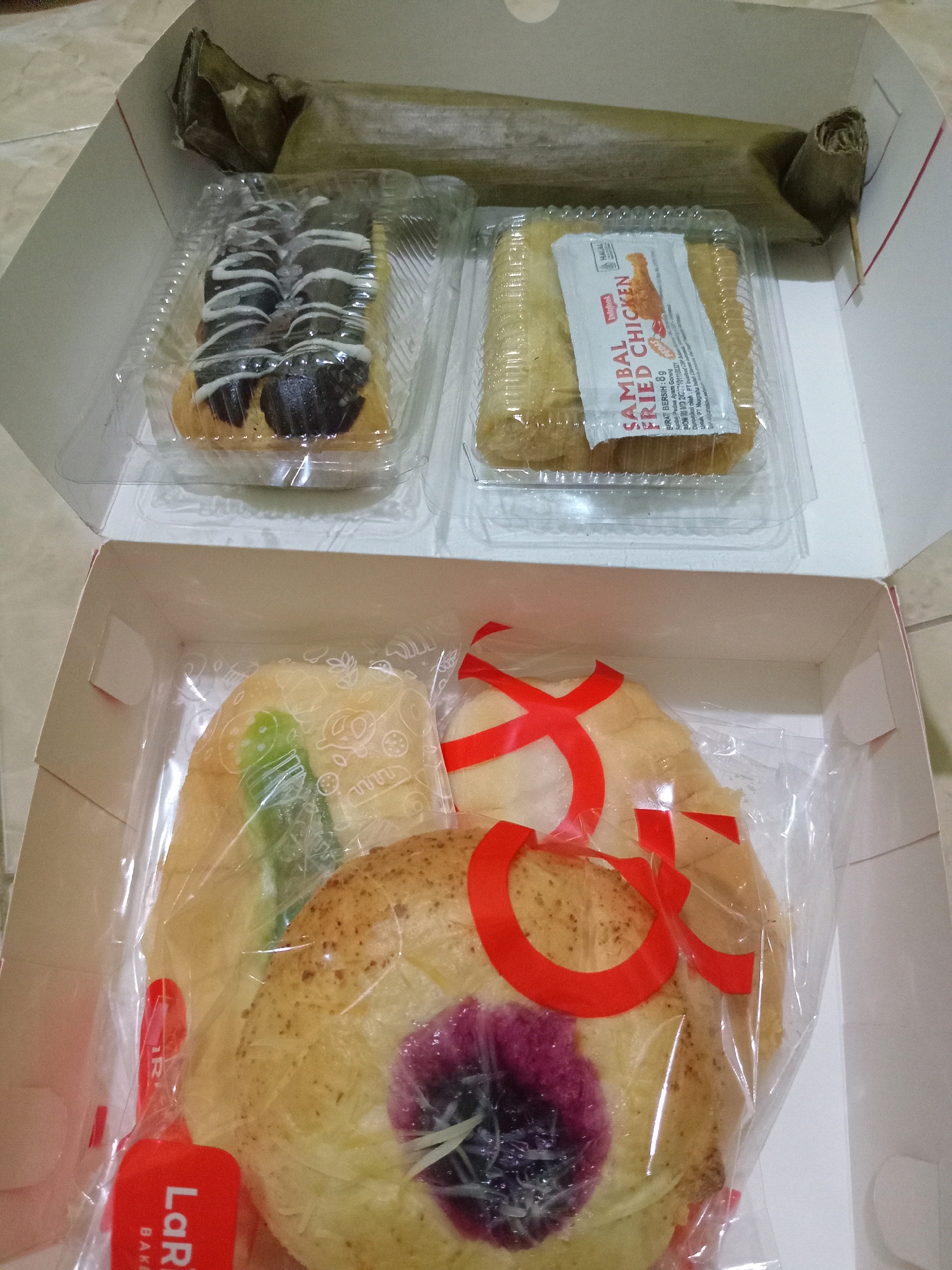 In Randuagung, Indonesia, a white cardboard box contains an assortment of pastries. The top section of the box holds two clear plastic containers. One container features two chocolate-frosted pastries with white icing drizzles. The second container holds a golden-brown pastry wrapped in clear plastic, with a small packet labeled "SAMBAL FRIED CHICKEN" placed on top. Behind these containers, a long, cylindrical item wrapped in a light green leaf material is visible. The bottom section of the box contains more pastries also wrapped in clear plastic. One pastry is a light brown bun with a purple filling and grated white cheese topping, adorned with a red ribbon. Another pastry, partially visible, is a light brown bun with a green filling. A red plastic tag with "LaR' BAKER" printed on it is attached to the packaging. The box is placed on a tiled floor.
