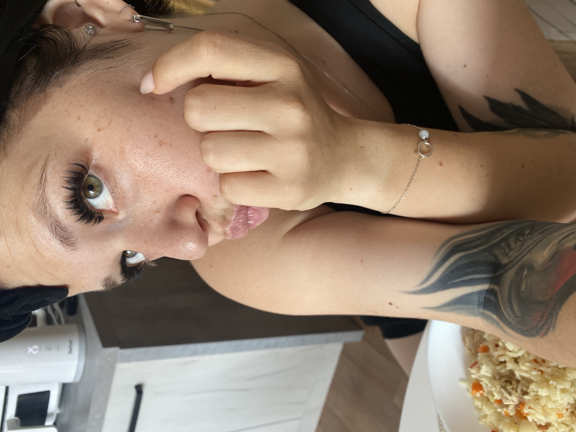 A close-up, slightly high-angle shot shows a woman with her face partially obscured by her hand, which is resting on her chin. She has light brown eyes, prominent eyelashes, and freckles scattered across her cheeks and nose. Her lips are slightly pursed. She is wearing a black tank top and a silver bracelet with two circular charms. A tattoo is visible on her left arm. In the background, out of focus, is a white countertop with kitchen appliances and cabinetry. A plate of rice with carrots and what appears to be chicken is on the left side of the frame. The scene is indoors, likely a kitchen or dining area, and there is no indication of it being in Saint Petersburg, Russia.
