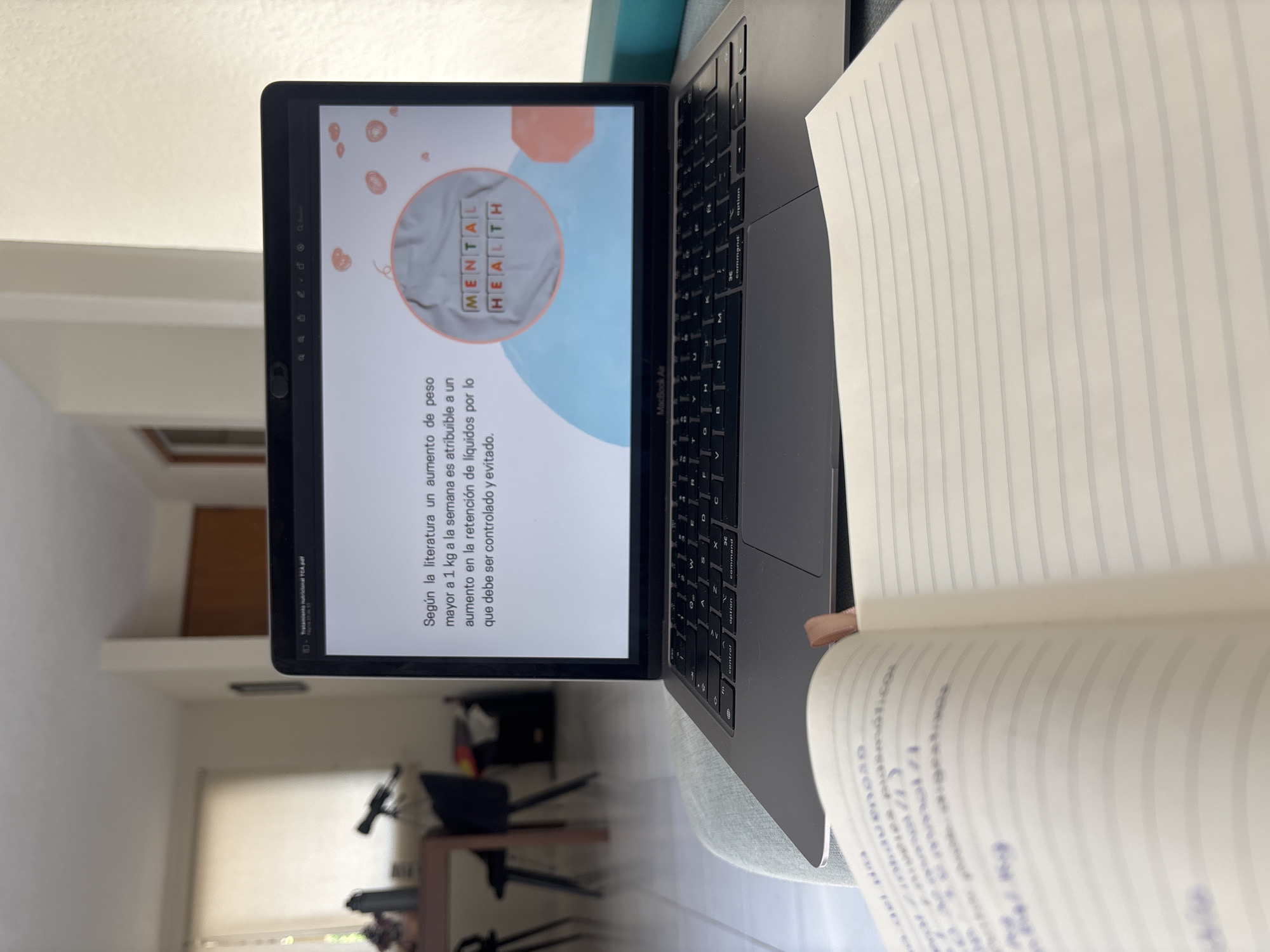 The image shows a laptop, likely a MacBook Air, open on what appears to be a sofa or chair. The screen displays a presentation slide with the words "MENTAL HEALTH" inside a circular graphic. Text in Spanish is visible below, discussing the literature suggesting that weight gain greater than 1 kg per week is attributable to fluid retention and should be monitored. A notebook or journal is also present, partly obscuring the right side of the laptop, with lined paper visible. The background suggests an interior setting, possibly a living room or study, with a window letting in natural light. There are some furniture and objects in the background, though they are partially out of focus. The image does not provide enough information to determine if it was taken in Puebla, Mexico, or any other specific location.