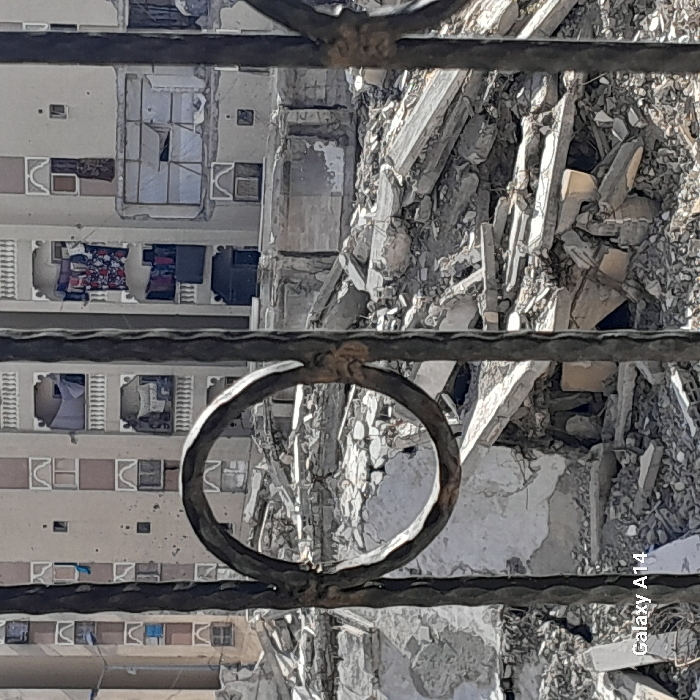 The image displays a scene of urban destruction in Khan Yunis, Palestinian Territory, viewed through dark, ornate metal bars in the foreground, one featuring a prominent circular design. The midground is dominated by an extensive field of concrete rubble, broken slabs, and shattered building debris, indicative of a demolished structure. In the background, a multi-story residential building stands partially damaged but intact, its light-colored facade exhibiting numerous broken windows, missing glass panes, and potential impact points. Several balconies on this background structure show items of clothing hanging, including patterned and solid fabrics. One balcony displays a banner with indistinct text and what appears to be a figure. The sky is clear. No individuals are visible within the frame.