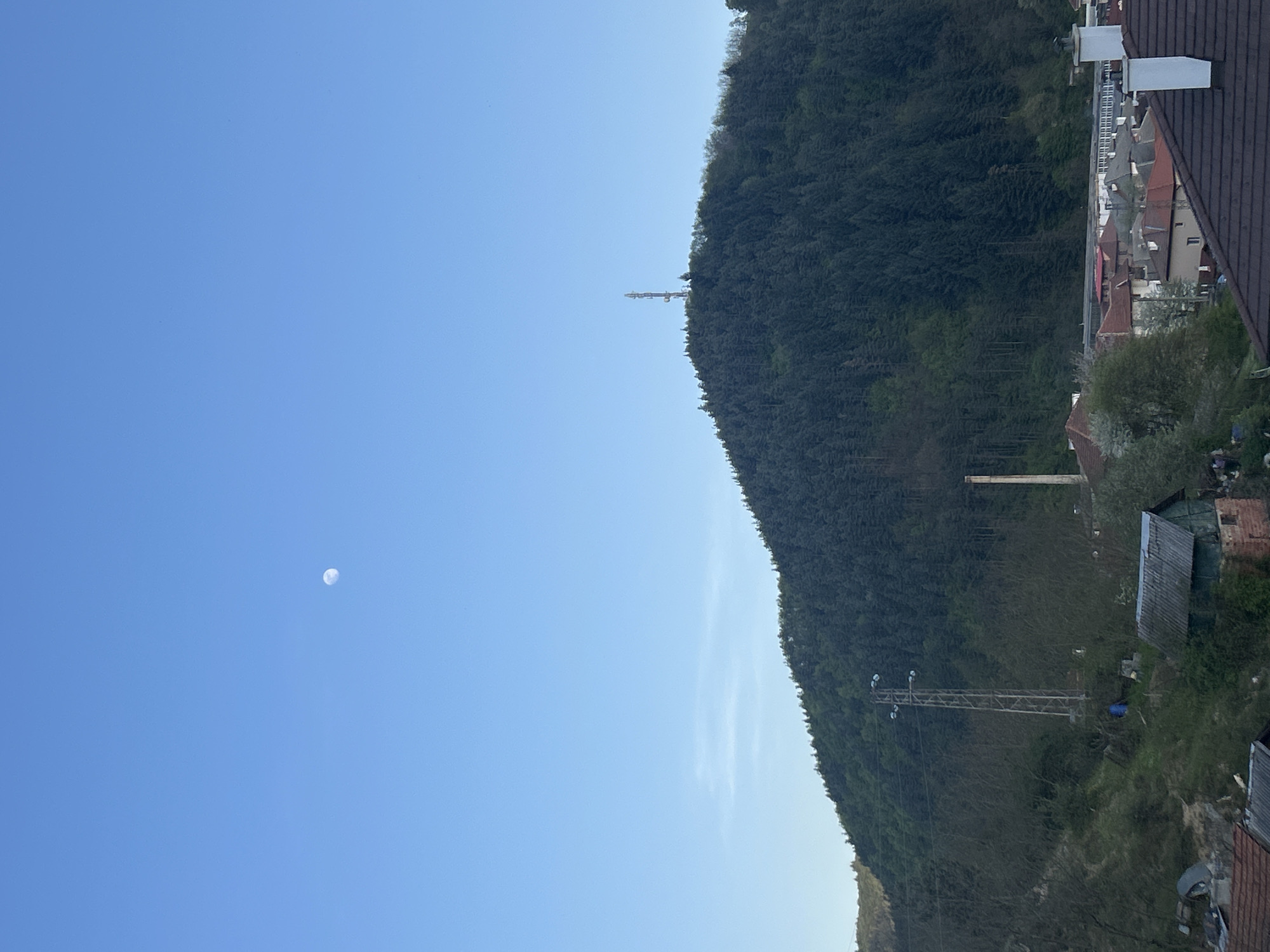 The image presents a tranquil, daytime landscape scene from Zlatograd, Bulgaria, encompassing a clear sky, a forested mountain, and a glimpse of a village.
The upper portion of the frame features a vast, bright blue sky, indicating clear weather. A distinct, white daytime moon is visible in the upper left quadrant of the sky. Below the sky, a large, verdant mountain or hill dominates the middle ground, densely covered with coniferous and deciduous trees, appearing lush and green. On the mountain's ridge, a slender communication tower or antenna stands tall against the sky. Further down the mountain slope, a power line structure, possibly a pylon or smaller transmission tower, is visible, partially integrated with the tree line.
In the bottom right corner of the image, parts of a village or town are visible, nestled at the base of the mountain. Several buildings with characteristic red and dark brown tiled roofs can be seen, suggesting traditional European, possibly Balkan, architecture. The scene is bathed in natural light, implying either early morning or late afternoon due to the gentle illumination and visible moon. The atmosphere conveyed is one of peace and natural beauty, with no human activity explicitly depicted. No visible text is present in the image.