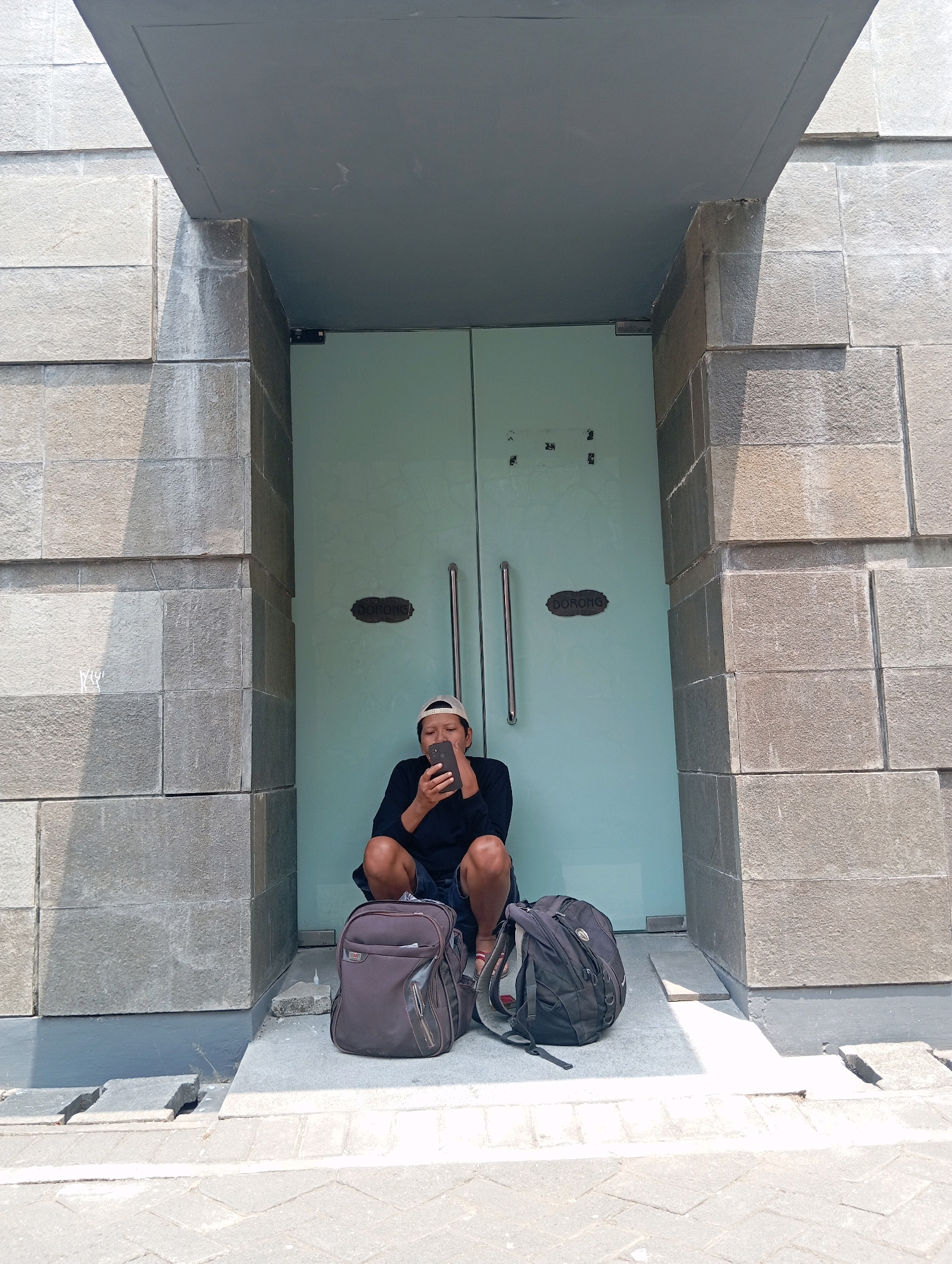 In the city of Blitar, Indonesia, a person wearing a black t-shirt, dark shorts, and a light-colored baseball cap is crouched in front of a large, pale blue glass door. They are looking down at a smartphone they are holding in their hands. Two backpacks are positioned on the ground in front of them, one a dark grey and the other black. The building's exterior is constructed with large, textured grey stone blocks. The scene appears to be an entrance or entryway to a building, possibly a commercial establishment or public building, with a covered overhang above. The ground in front of the door is paved with light-colored stones.
