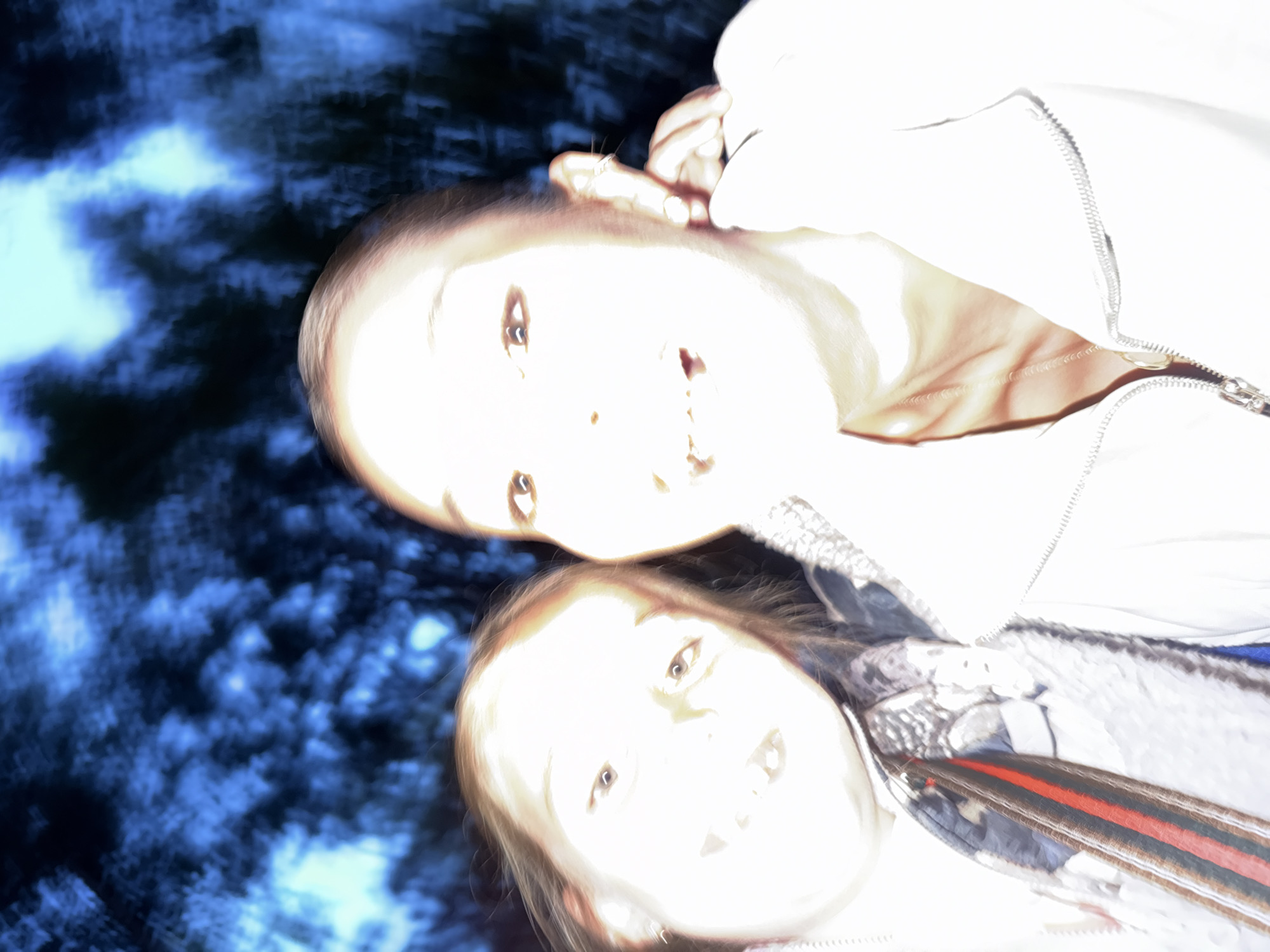 This is a blurry, overexposed selfie of two females, likely taken outdoors at night, indicated by the dark, out-of-focus background resembling trees illuminated by a bright light source. The two individuals are close together, with one positioned slightly in front of the other. Both are looking towards the camera. The person in the foreground on the left appears to be a child with brown hair, wearing a scarf with red and black stripes. The person on the right is an adult with brown hair, wearing a white zippered jacket. Their facial features are indistinct due to the blur and overexposure. The overall impression is of a casual, candid moment captured in low light conditions. No specific landmarks or context are discernible to place it within Krasnoyarsk, Russia, or any other location.