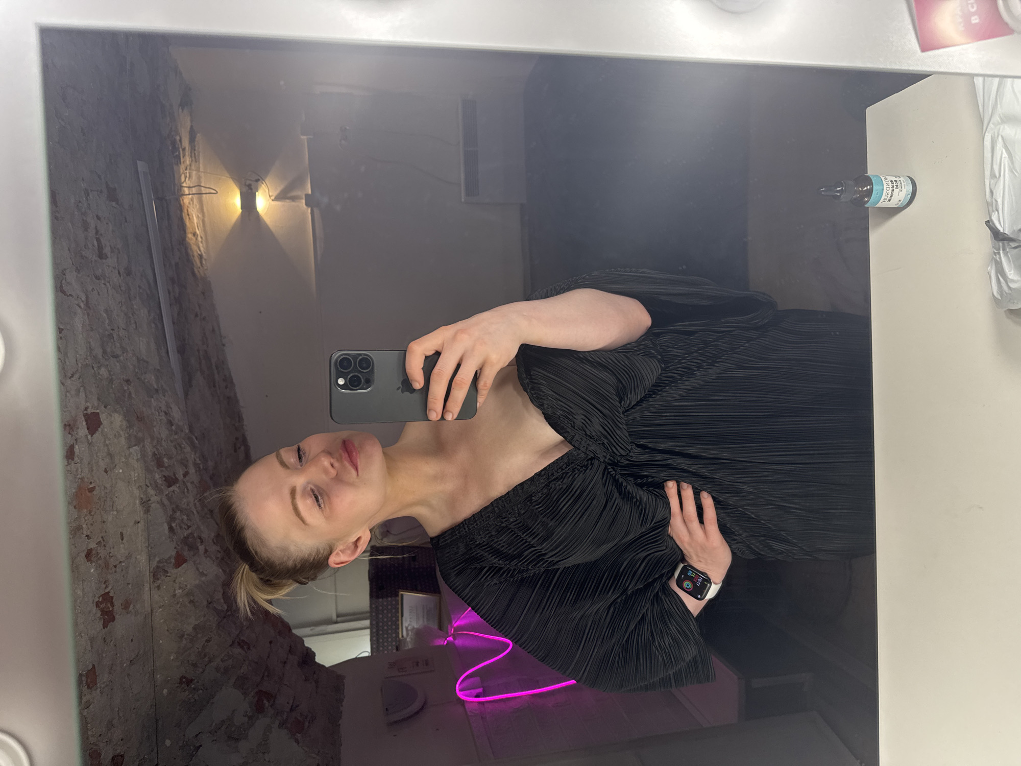 A woman is taking a selfie in a mirror. She's wearing a black dress with a pleated texture and an Apple Watch on her left wrist. The background reflected in the mirror shows a brick wall and a neon pink light. On the counter next to the mirror, there is a small bottle and some other items partially visible. The mirror is surrounded by a frame with lights. The location is Saint Petersburg, Russia.